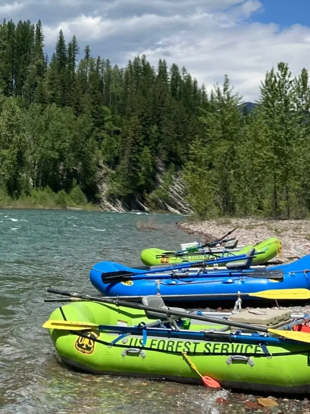 Multiple center frame oar rafts in bright colors on a Western Montana riverbank with a forest and mountains in the background.