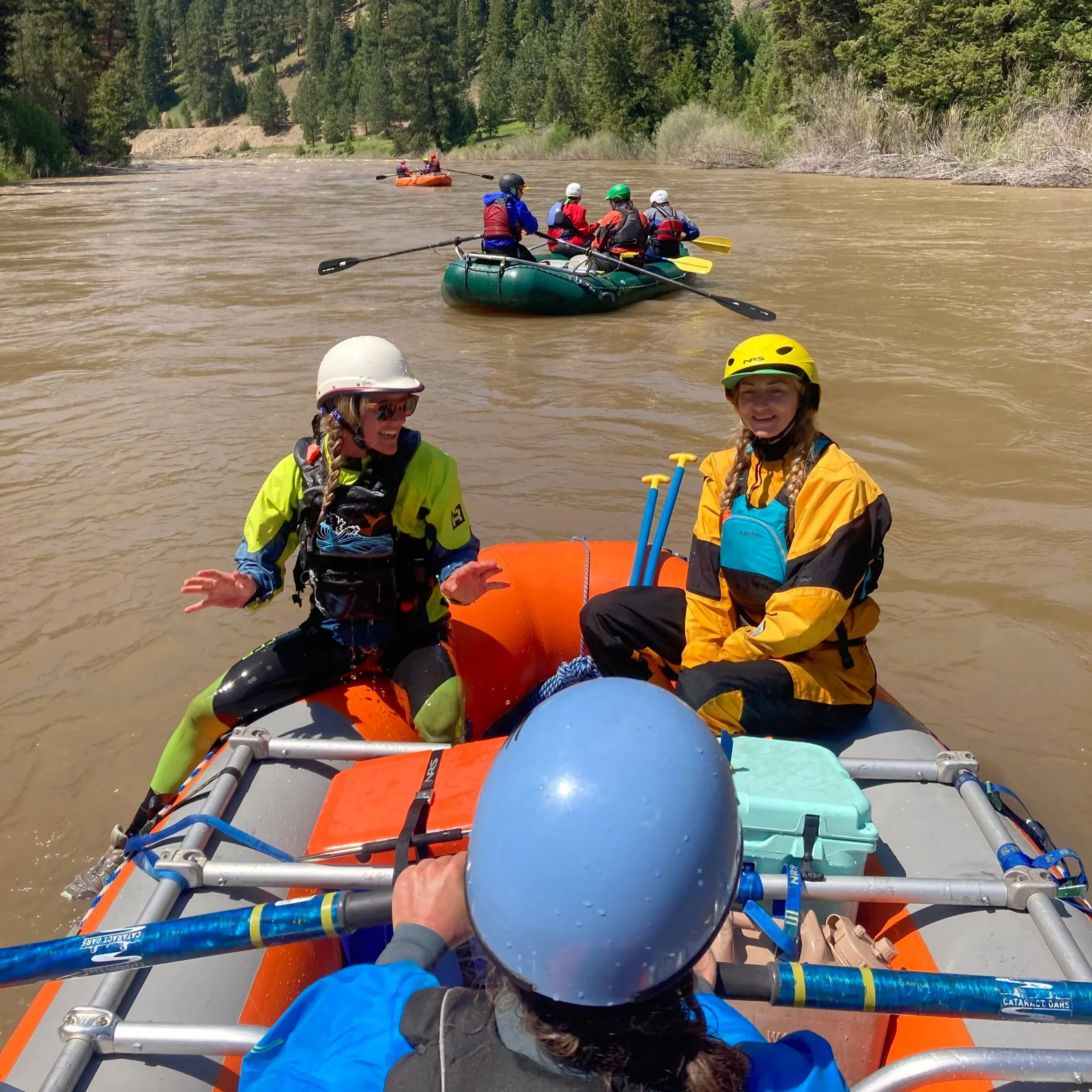 Women are rowing a rafting on a Montana river. Two women in safety gear and helmets are sitting on the front part of an inflatable raft, smiling and talking. Other rafts with groups of people are visible in the background.