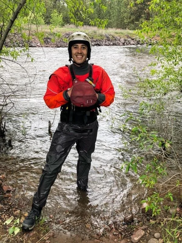 A man dressed in outdoor gear, wearing a helmet and waterproof pants, standing in shallow water near a river, holding a small bag and smiling.