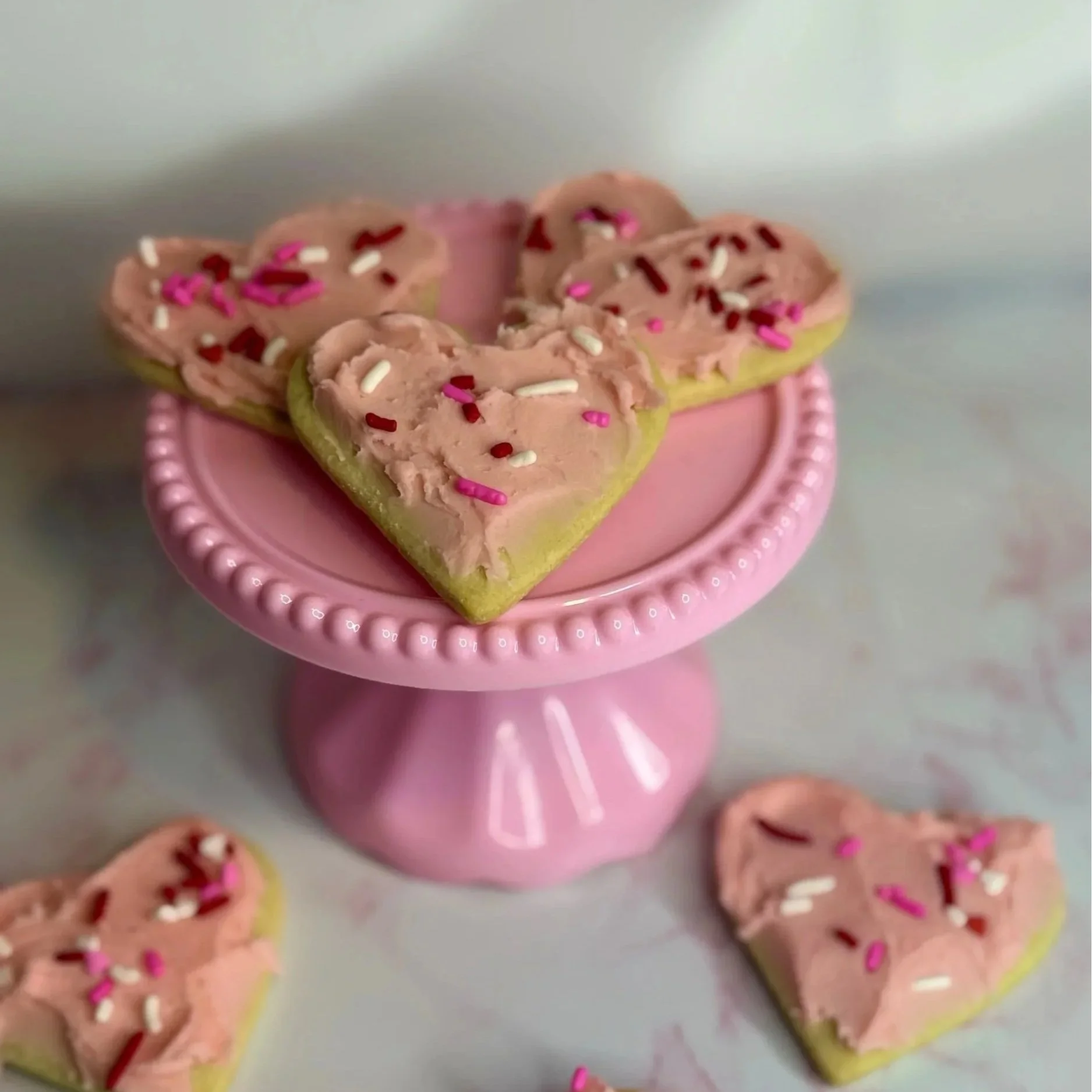 Heart-shaped cookies with pink frosting and multicolored sprinkles on a pink cake stand.