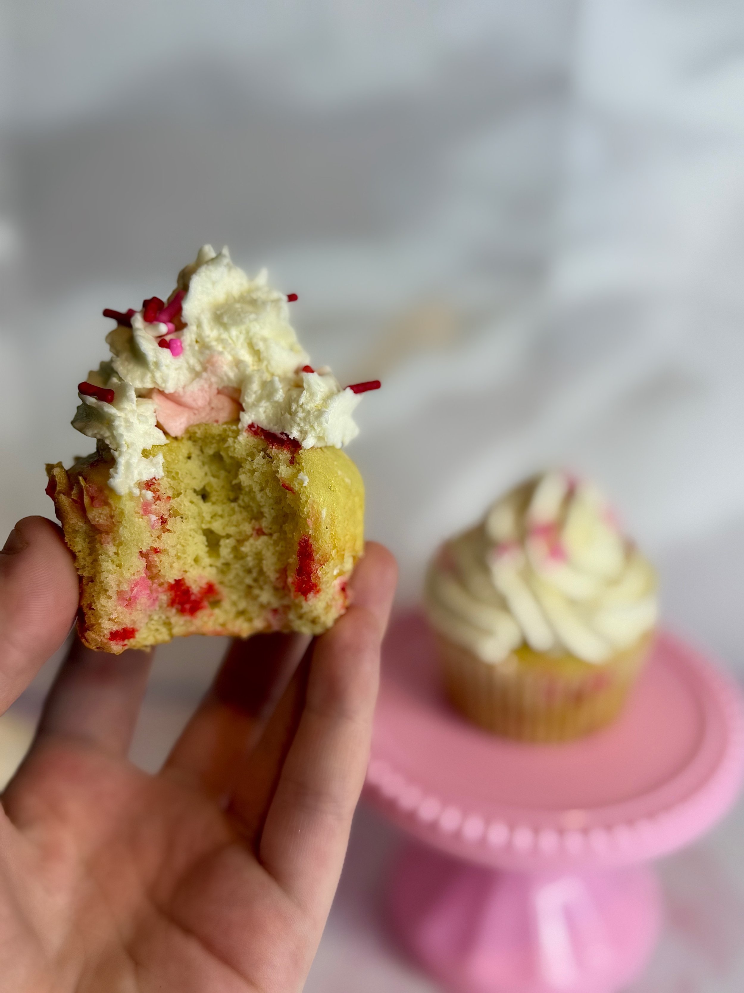 A hand holding a bitten cupcake with white frosting and pink sprinkles, with another whole cupcake on a pink cake stand in the background.