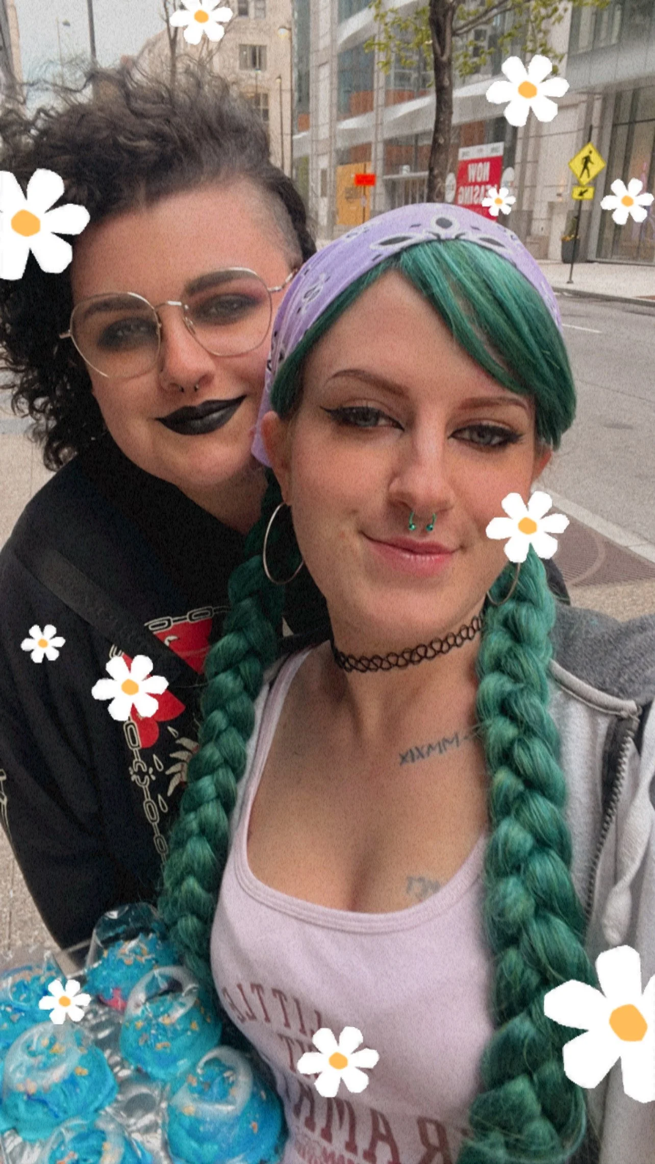 Two women taking a selfie outdoors with a city street background. One has green braided hair, a purple bandana, and septum piercings, while the other has short curly hair, glasses, and dark lipstick. The photo has a floral filter overlay.