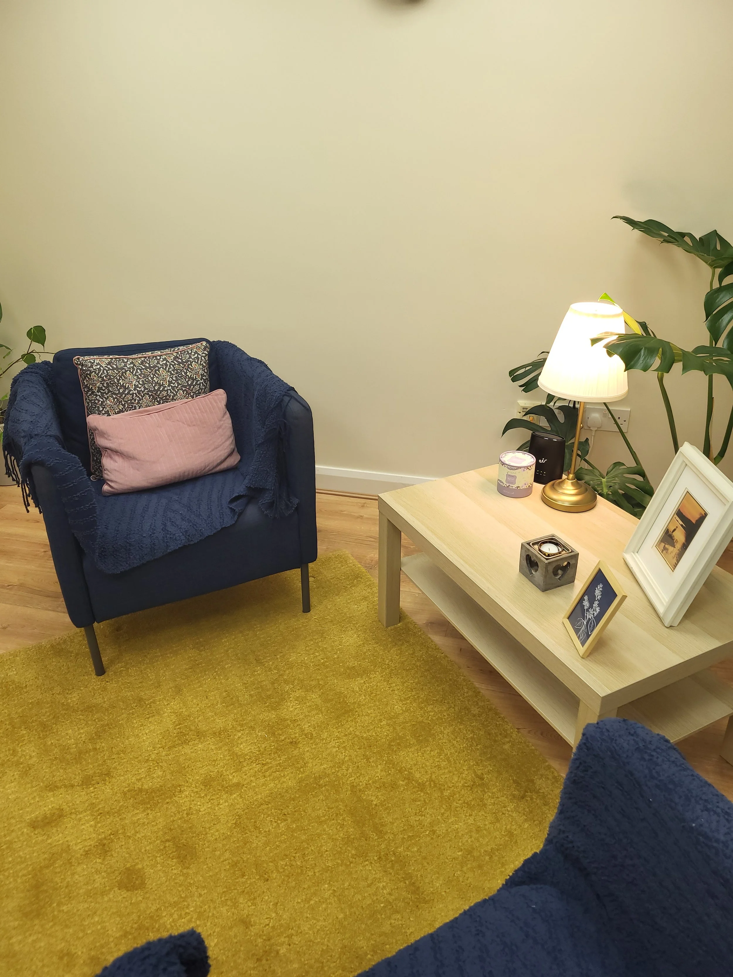 A cozy living room corner with a blue armchair, a light wood coffee table, and a mustard yellow rug. The table has a white framed photo, a small black speaker, a candle, and a small decorative box. A white lamp with a beige shade and some large green plants are in the background.