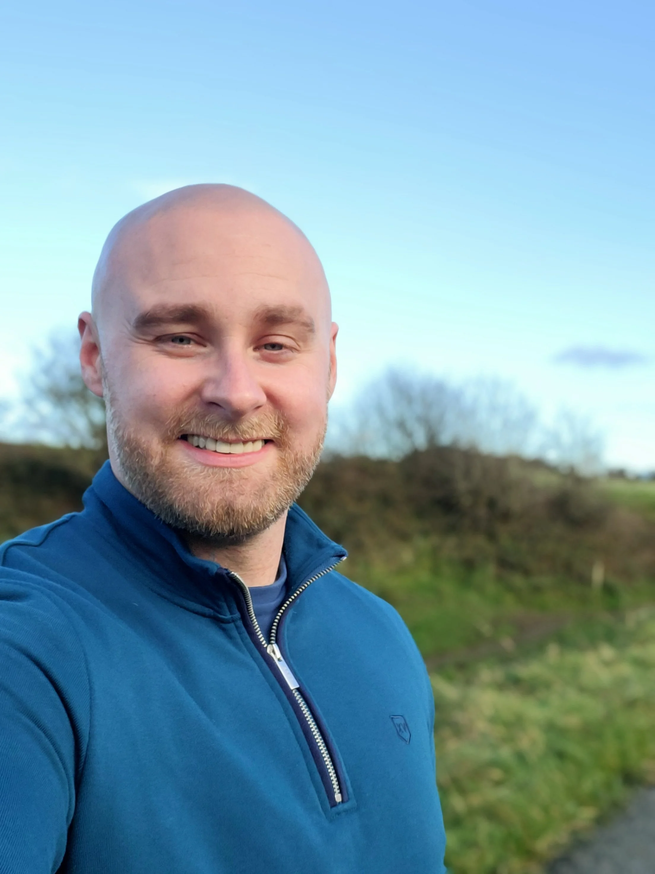 A smiling man with a bald head and beard wearing a blue zip-up jacket outdoors on a clear day with trees and a grassy area in the background. A profile picture for a psychotherapy & Counselling practice.