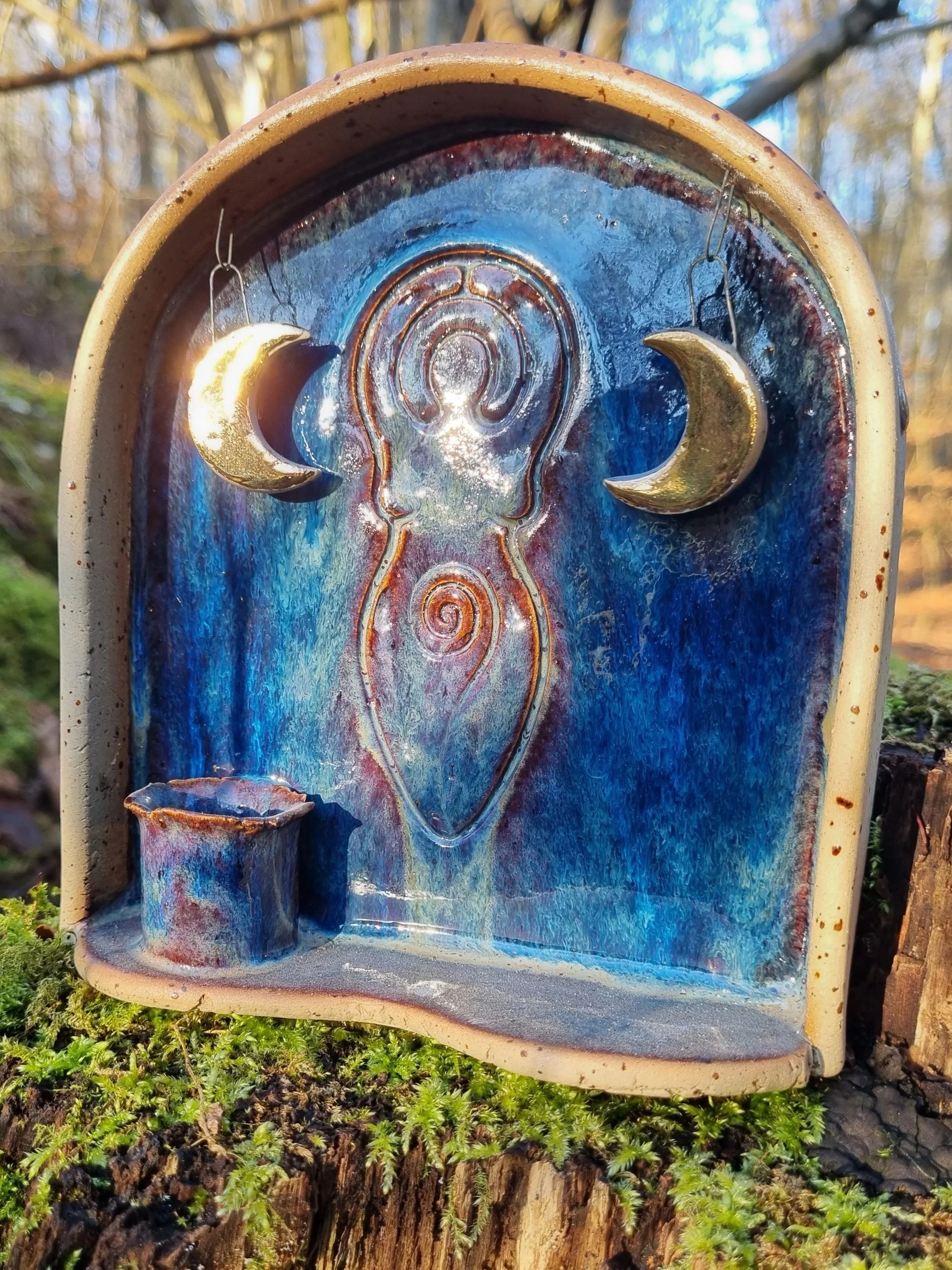 Decorative ceramic water fountain with moon and sun symbols, featuring gold crescent moons, a sun face, and a small basin, set outdoors on mossy ground.