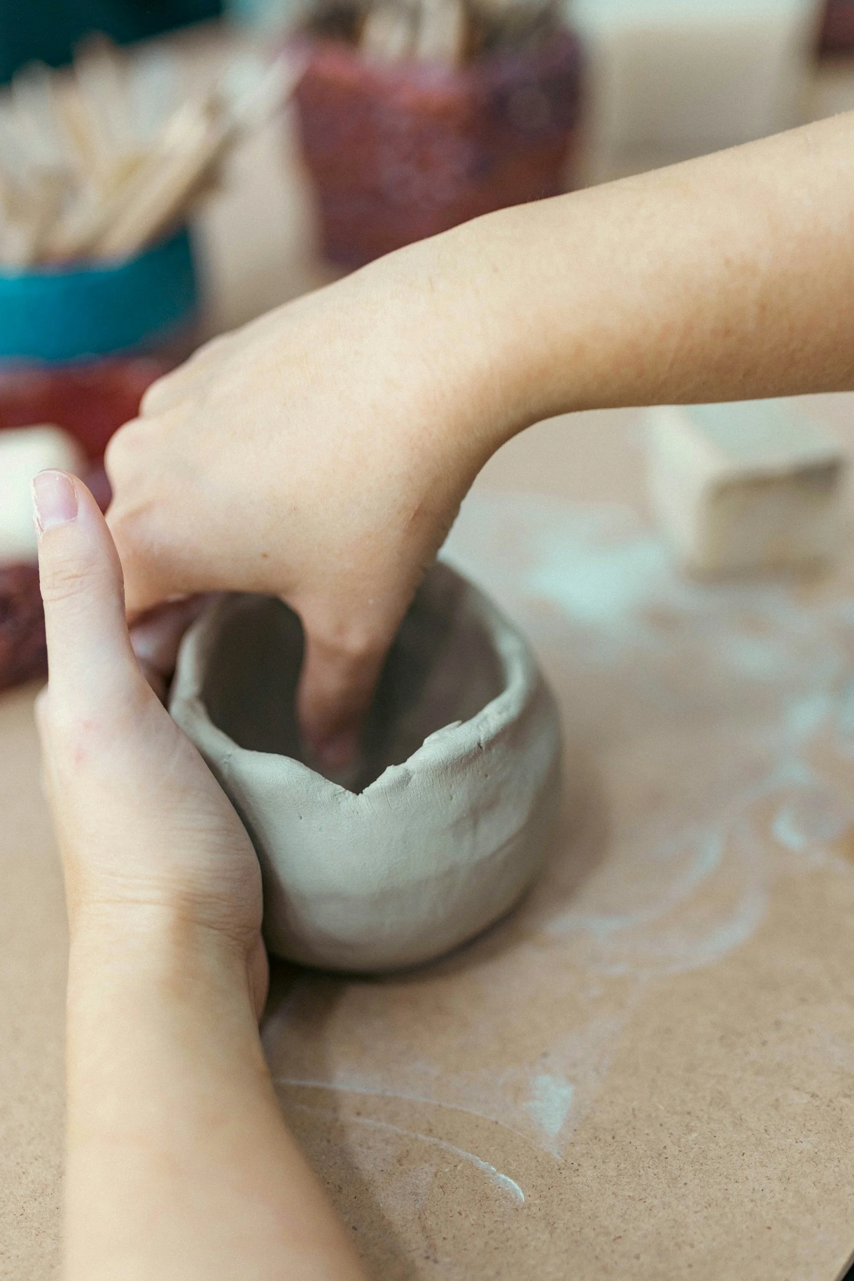 A person is shaping a small clay bowl with their hands, working on a pottery wheel or work surface. There are pottery tools and other clay pieces in the blurred background.