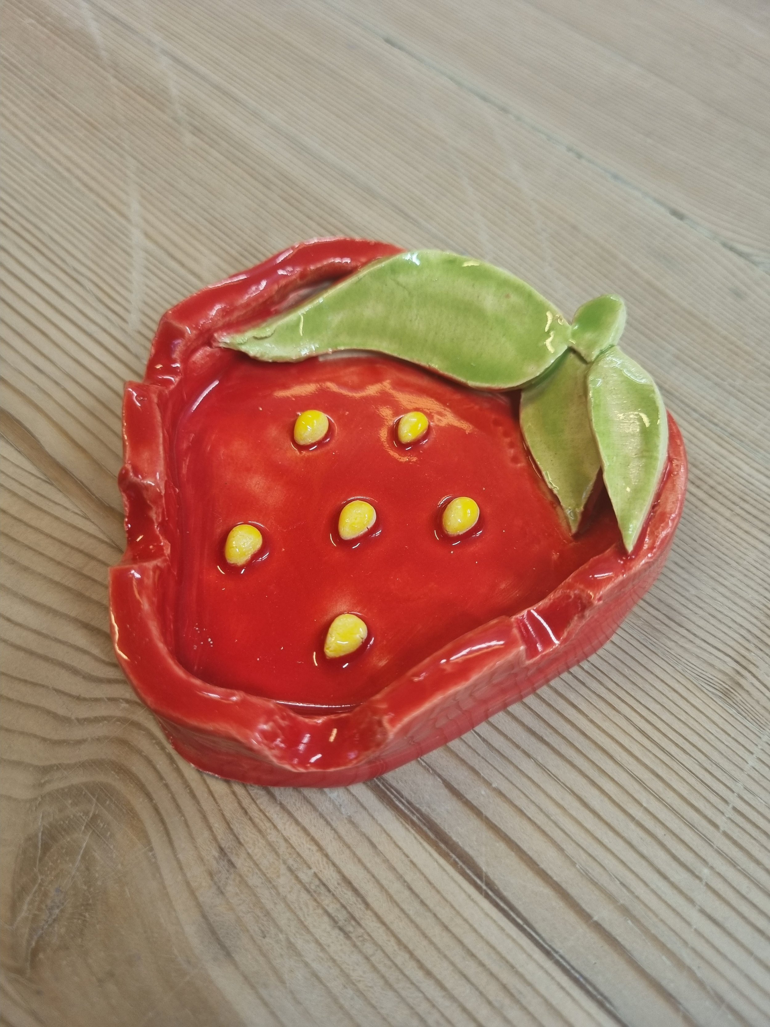 A decorative ceramic bowl shaped like a red strawberry with green leaves, filled with yellow seeds and placed on a wooden surface.