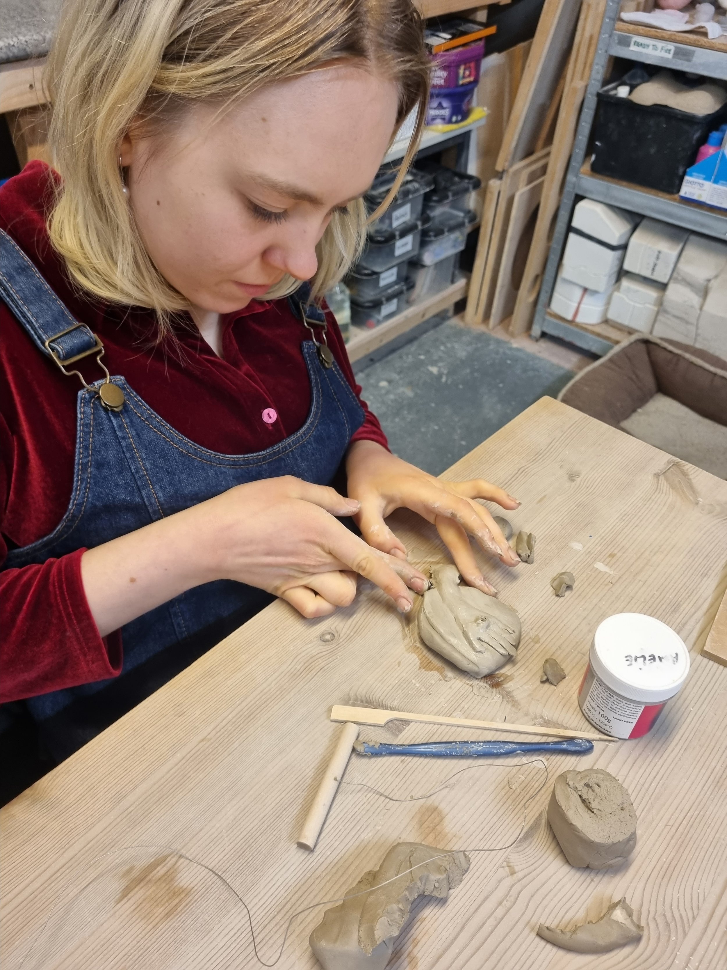 A woman with blonde hair wearing a red shirt and denim apron working on a piece of pottery at a wooden table in a pottery studio, surrounded by pottery tools and clay pieces.