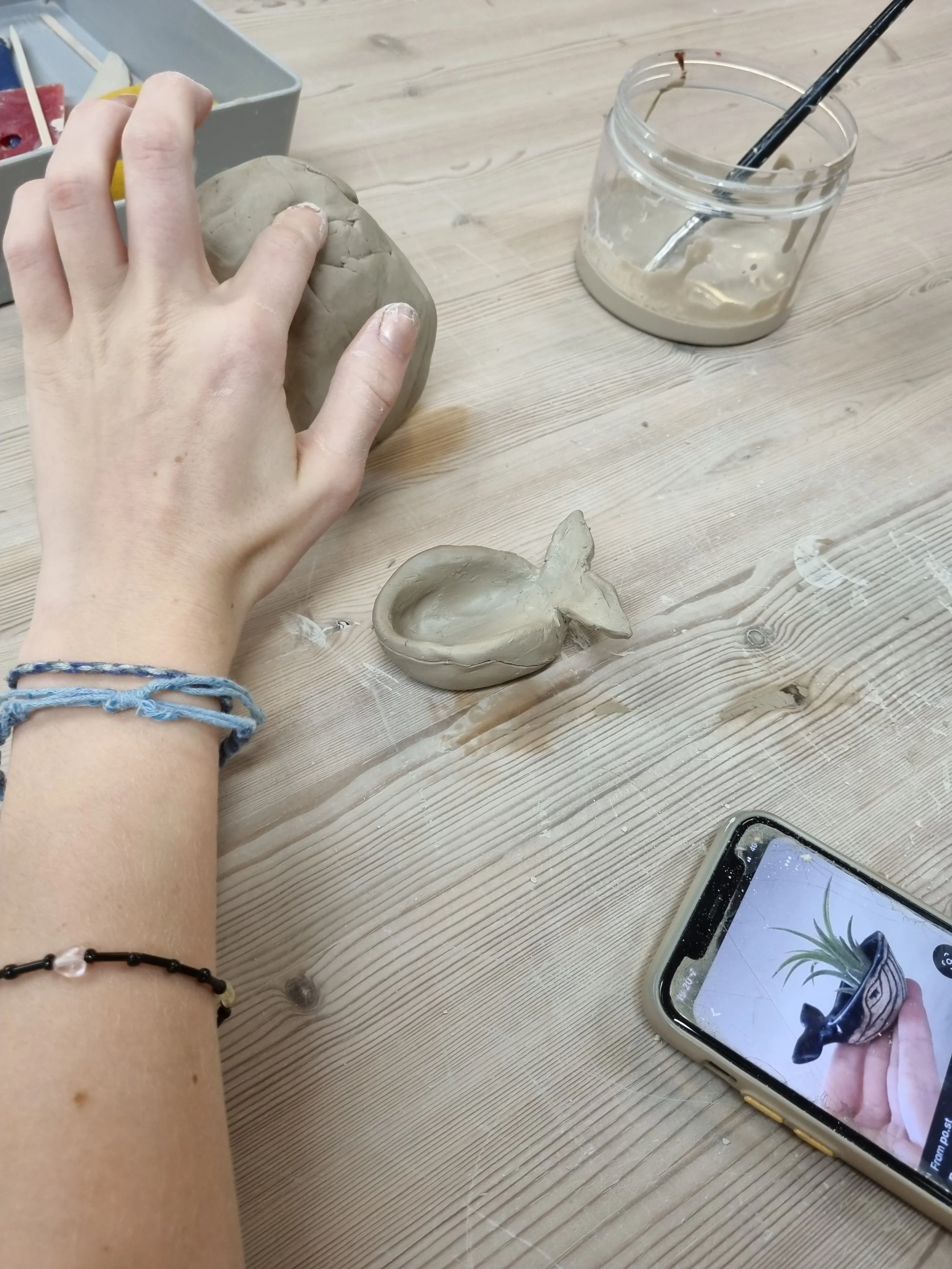 Someone shaping gray clay into a small bowl on a wooden table with a hand, a container with more clay and tools, a smartphone with a plant photo, and a glass jar with remaining clay and a brush inside.
