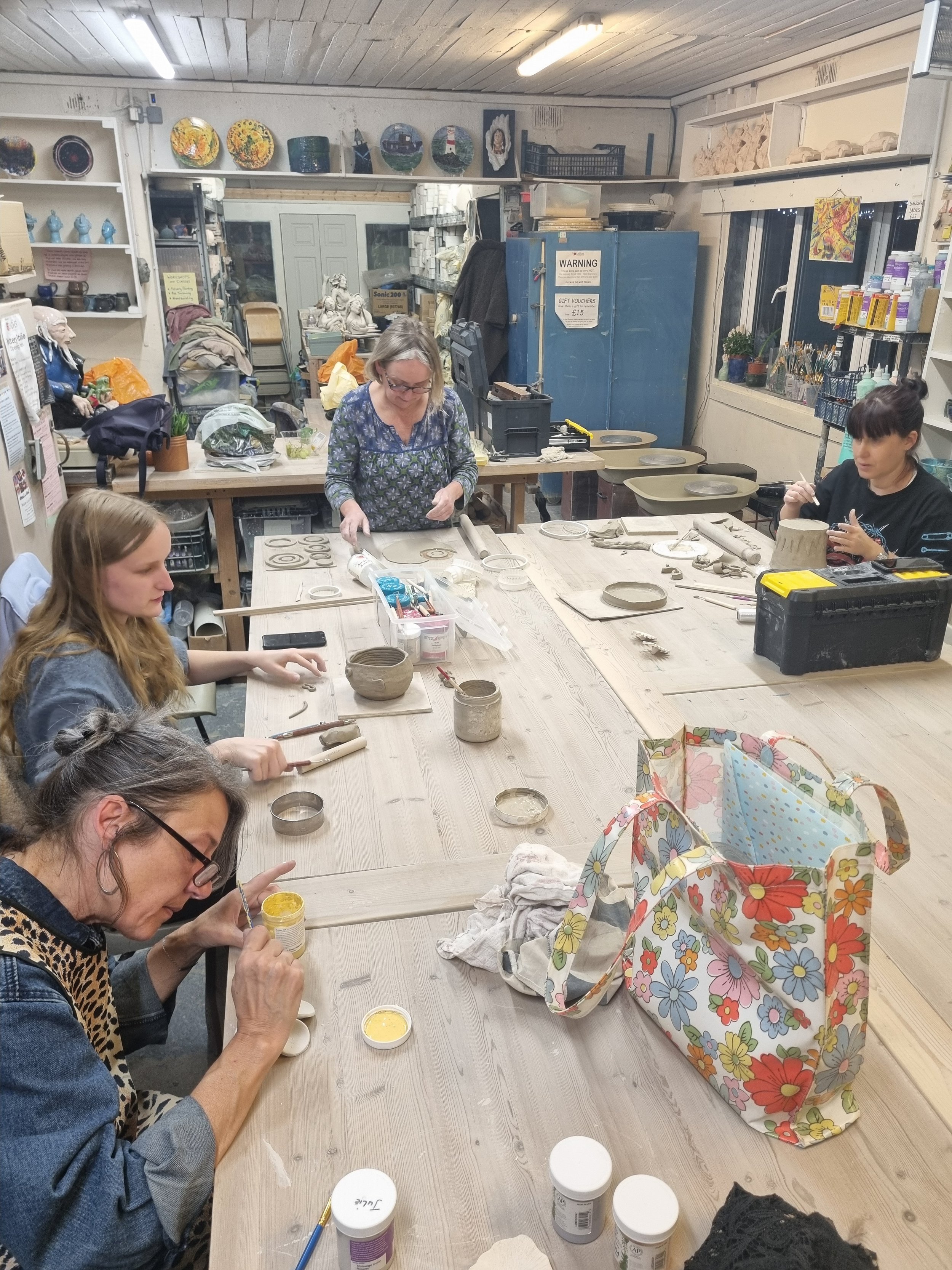 People participating in a pottery class, working on ceramic pieces at a large table in a studio filled with art supplies and decorated ceramics.