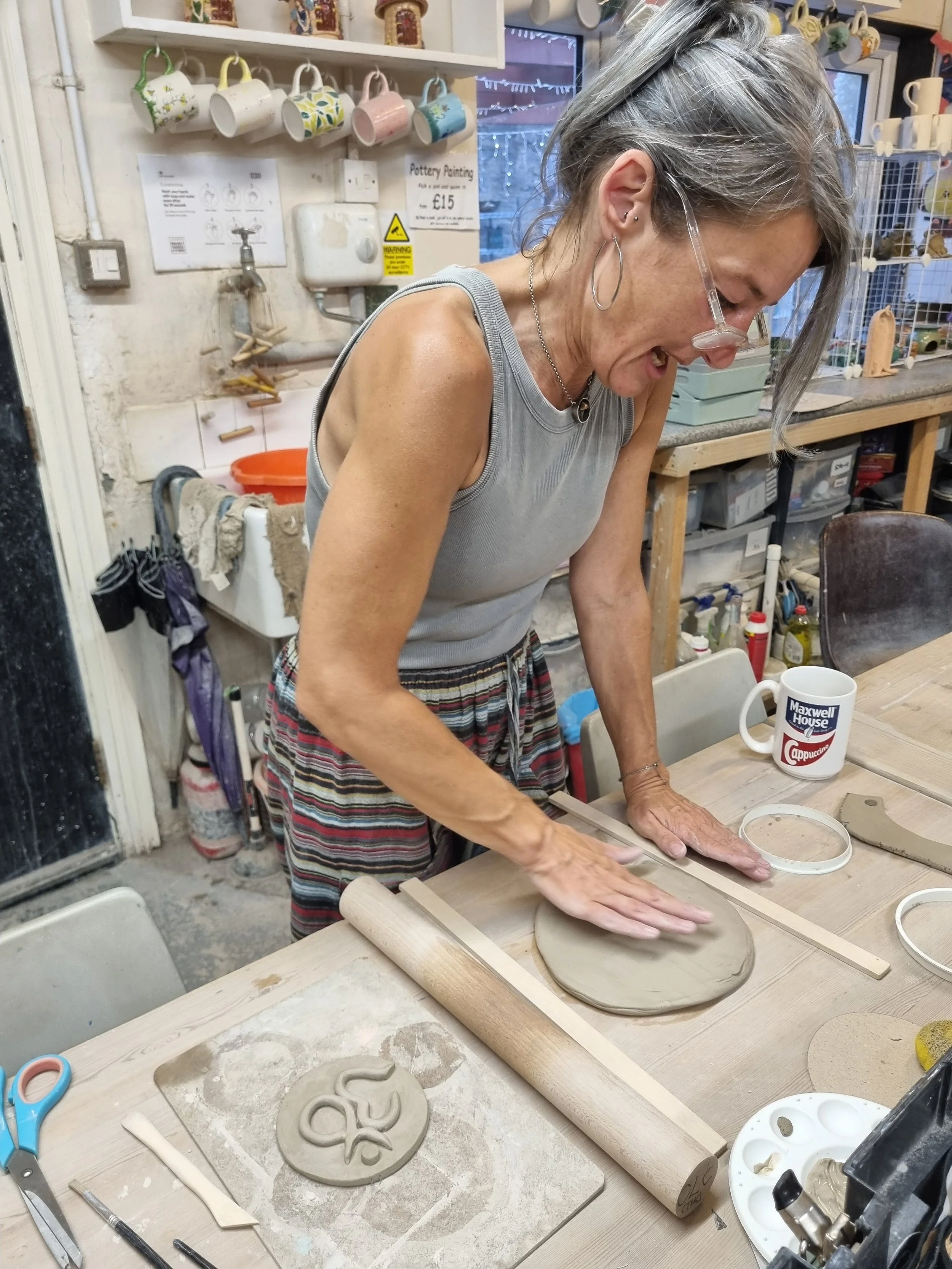 A woman working on a pottery wheel, shaping a clay plate in a pottery studio with various ceramics, tools, and supplies around her.