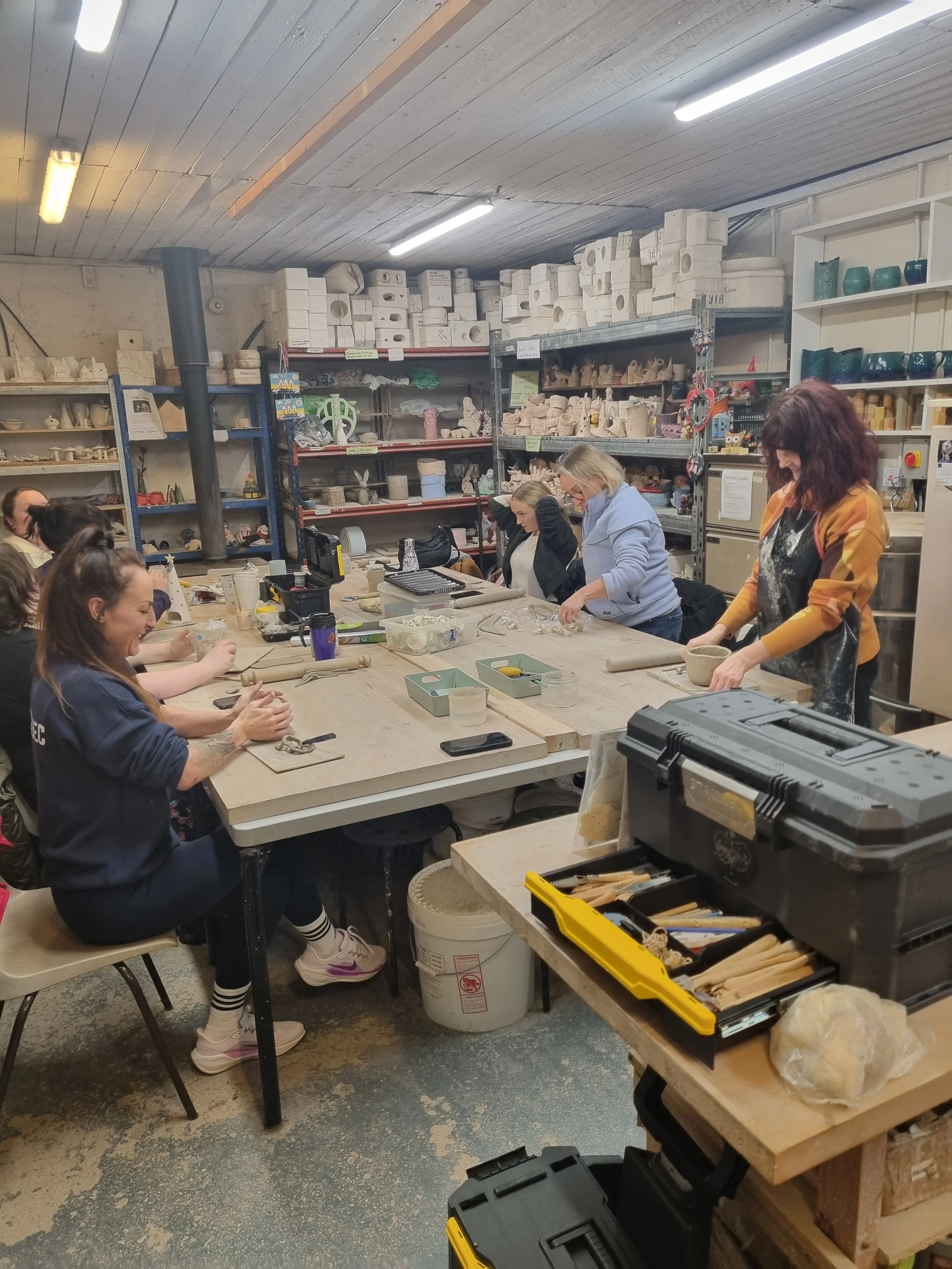 People gathered around a table in a pottery workshop, working on ceramic projects with various tools and clay pieces.