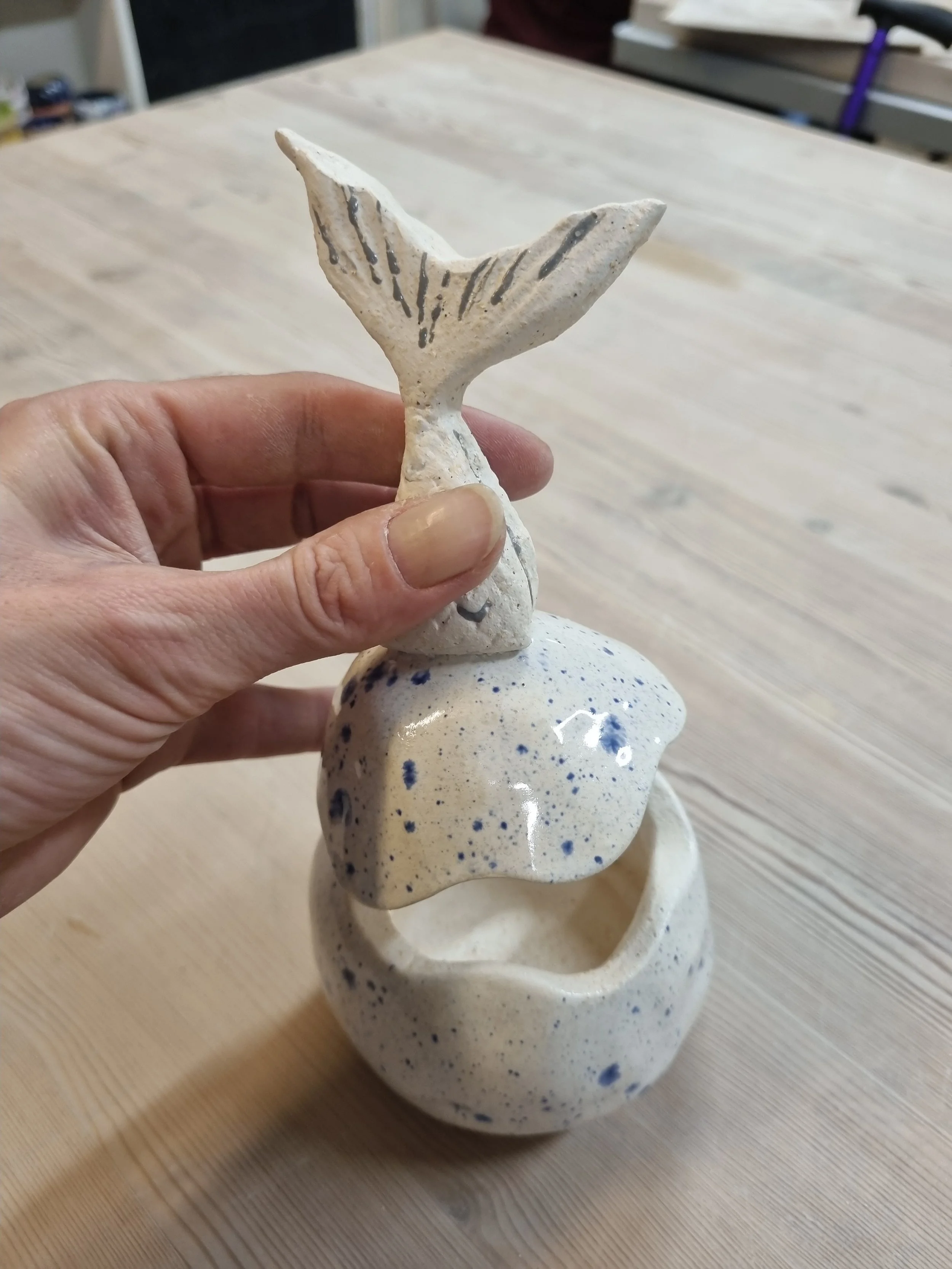 Hand holding ceramic sculpture of a whale tail on top of stacked, rounded ceramic pieces with blue speckles.