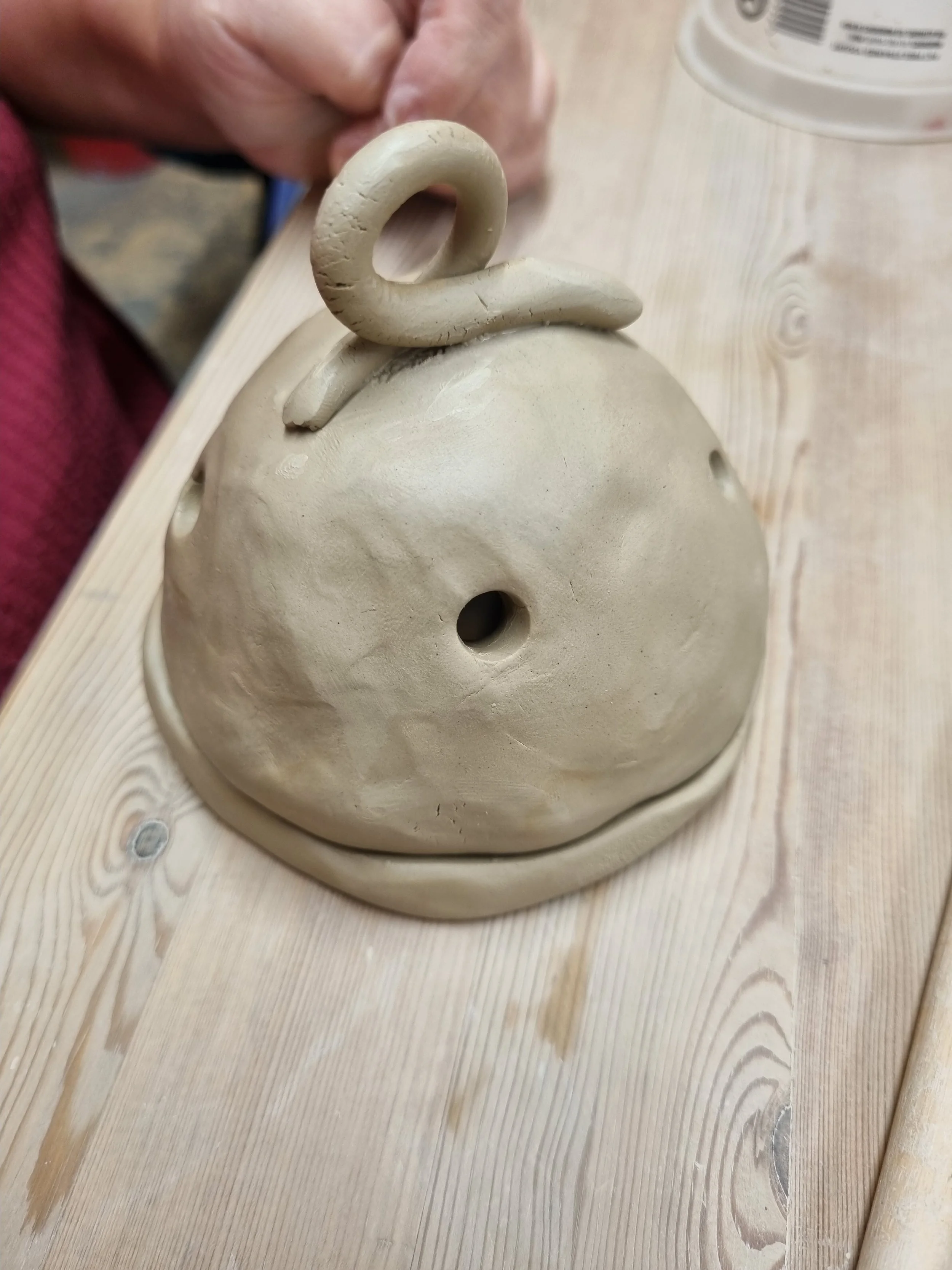 Unfinished pottery piece of a round object with a hole in the center, shaped like a kettle or teapot, on a wooden table.