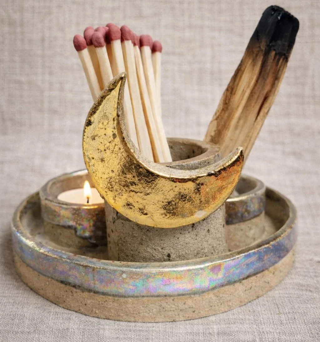 A decorative candle holder with a burning candle, containing a gold crescent moon, a bundle of matches, and a piece of wood, set on a circular base with a textured surface.