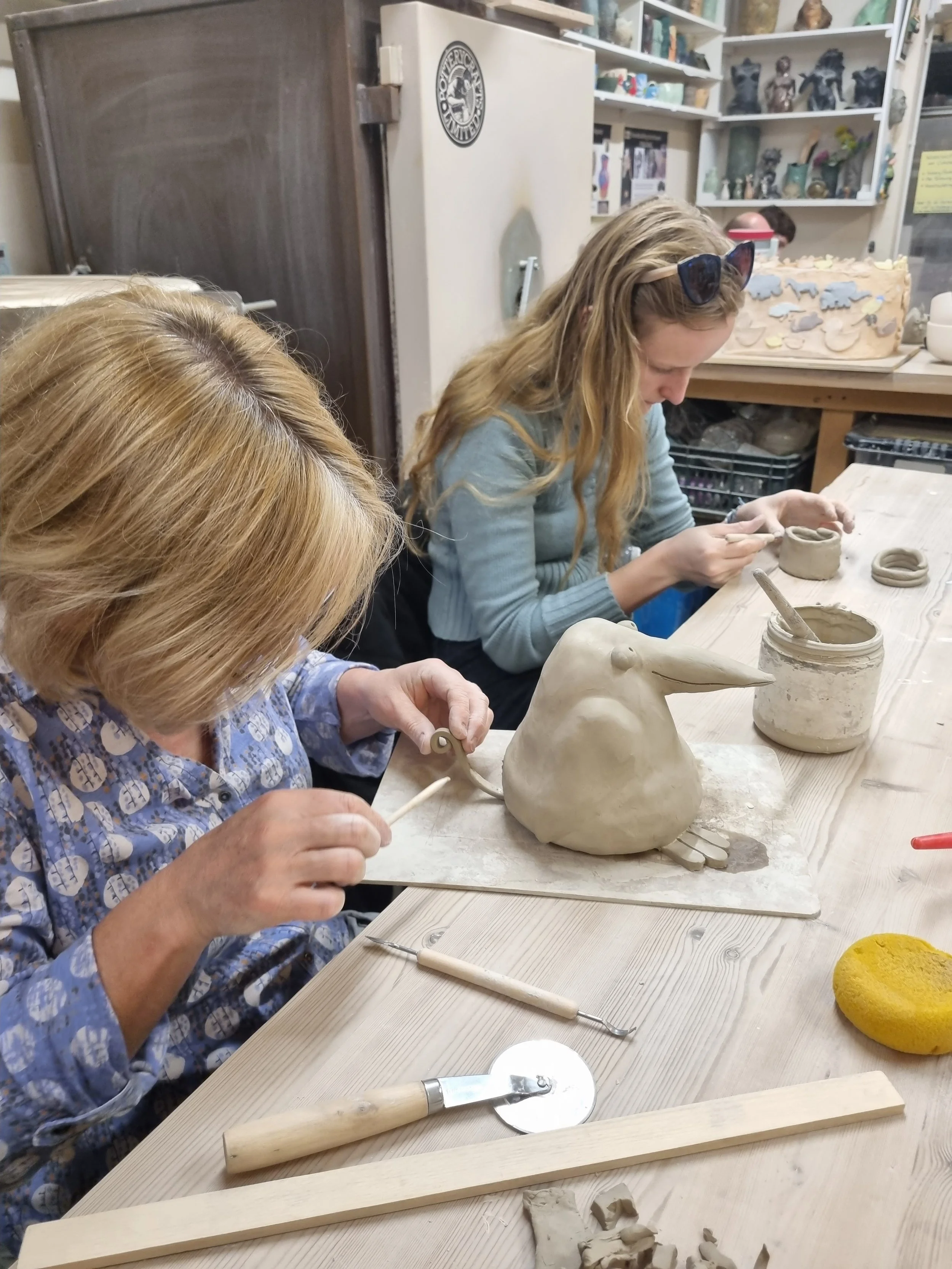 Two women working on ceramic sculptures at a pottery studio, including a bird sculpture, with various tools and clay on the table.