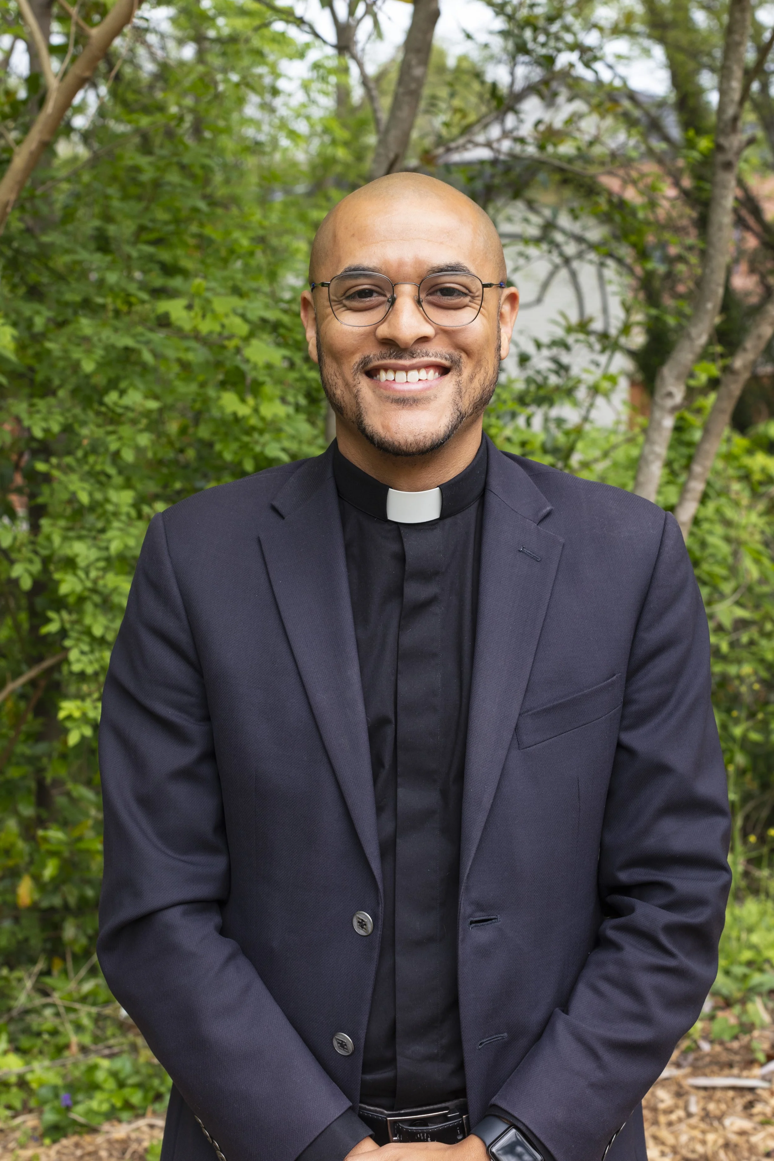 A smiling man with glasses, dressed in a black suit and clergy collar, standing outdoors among green trees.