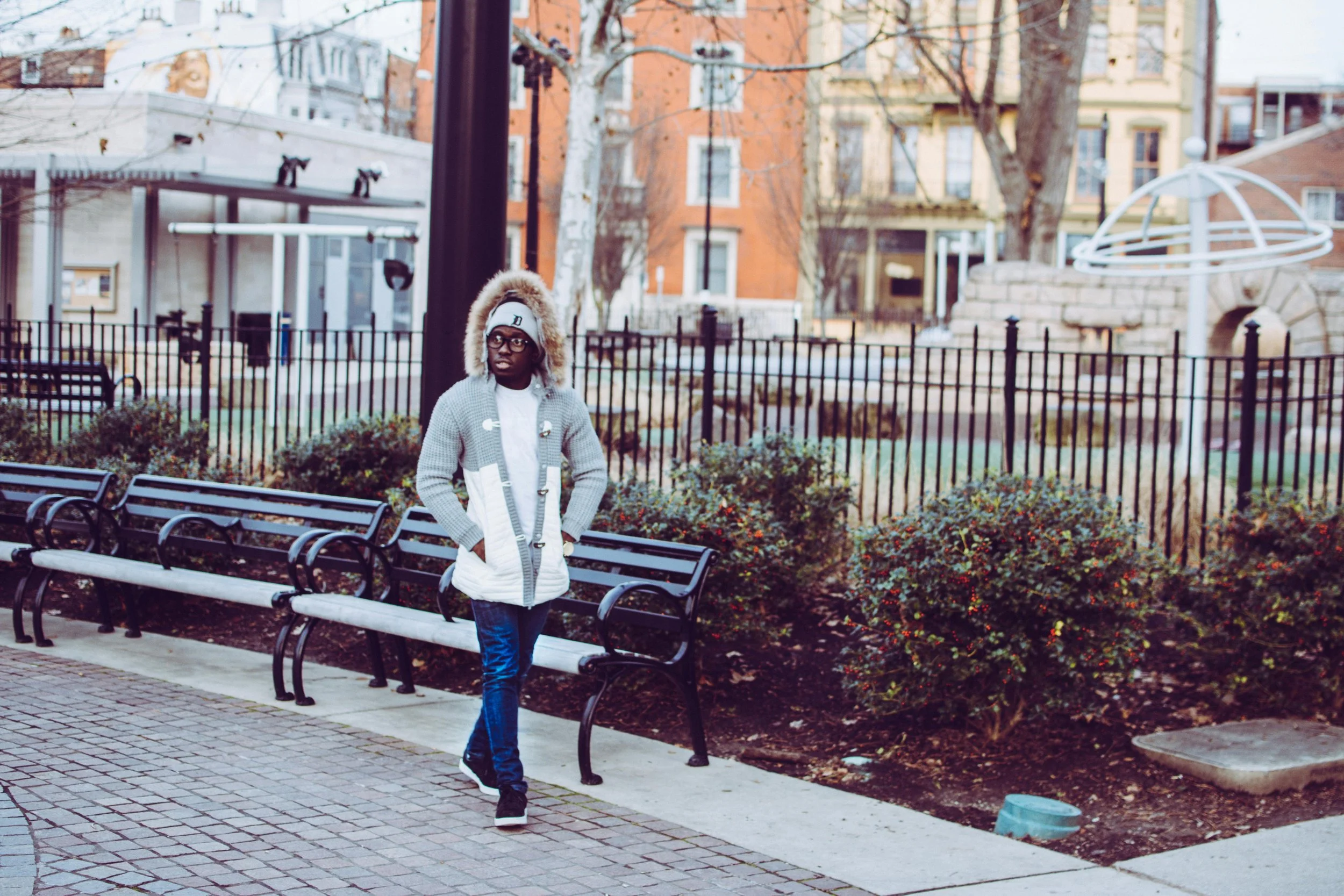 A young man walking in a park, wearing a gray coat with a fur hood, black glasses, and a white cap, with benches and bushes in the background.