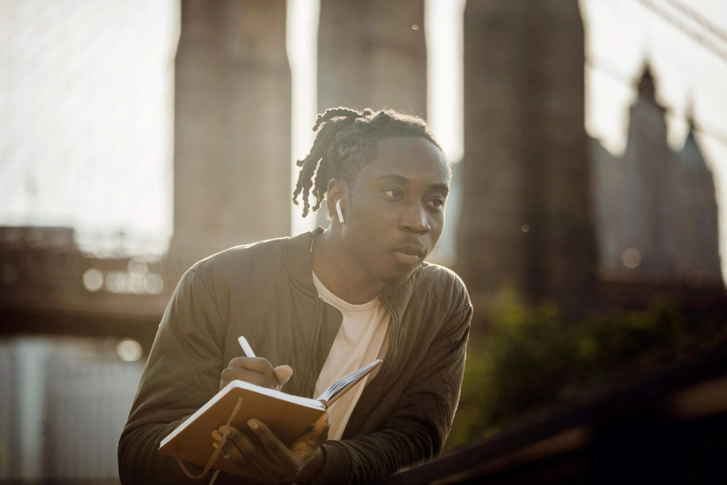 A young man with dreadlocks wearing a brown jacket and white shirt, sitting indoors with sunlight streaming through large windows, writing in a notebook, wearing wireless earbuds.