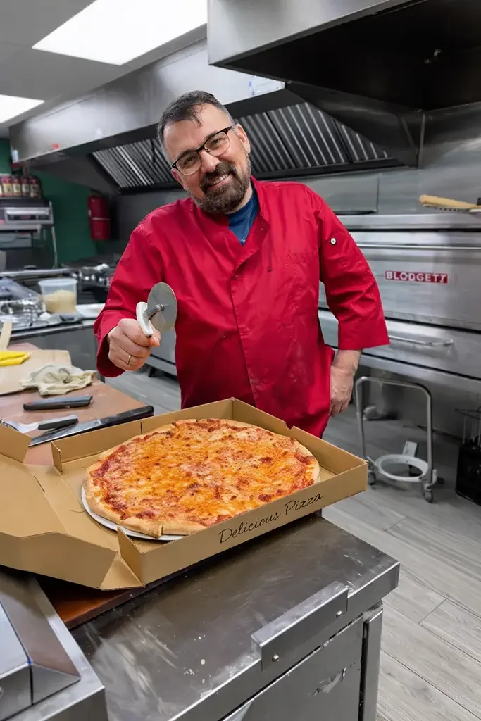 A man wearing a red chef coat and glasses standing in a commercial kitchen, holding a pizza cutter next to a large cheese pizza in a cardboard box.