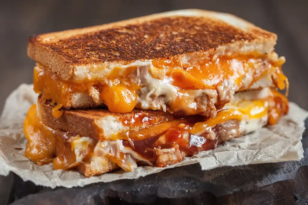 Close-up of a grilled cheese sandwich with melted cheese, eggs, and possibly chicken on parchment paper.