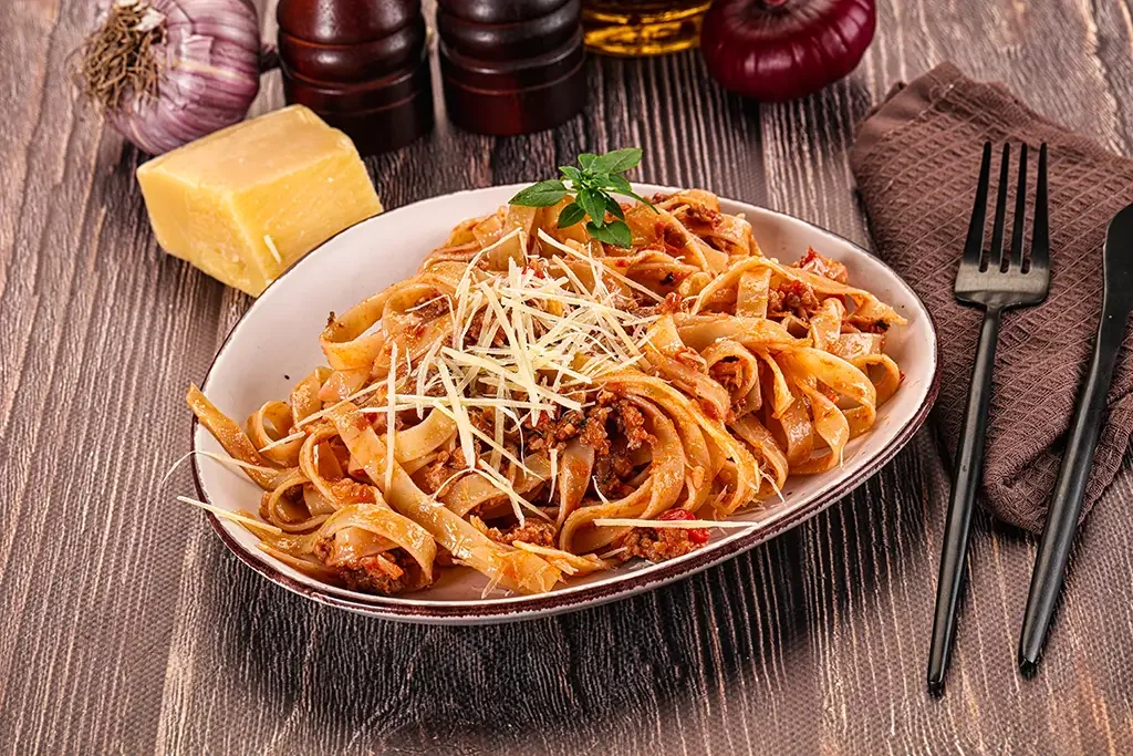 Plate of fettuccine pasta with meat sauce, topped with shredded cheese and a basil garnish. A block of Parmesan cheese, garlic, and condiments are on the table. A napkin, fork, and knife are beside the plate.