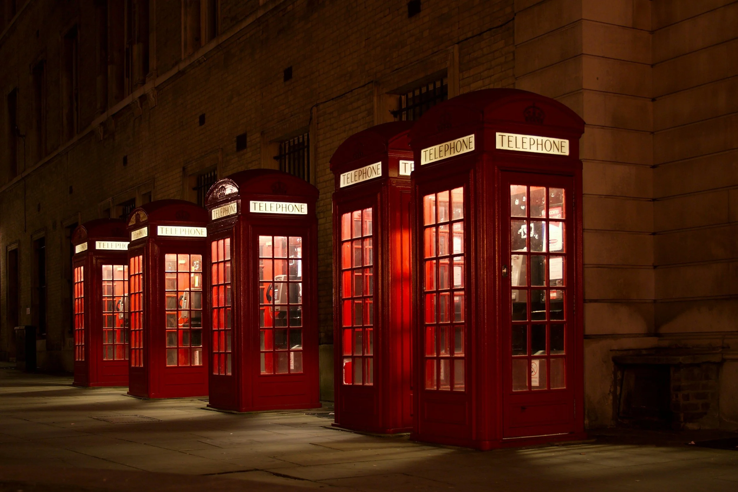Four traditional red British telephone booths illuminated at night, lined up along a city sidewalk against a brick building.