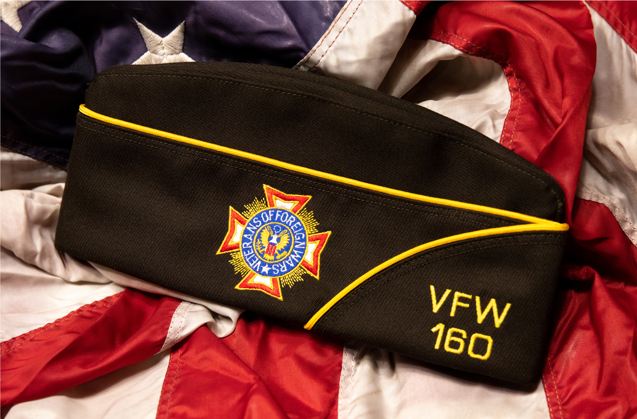 A black firefighter's hat with yellow piping and an embroidered emblem that reads 'Veterans of Foreign Wars' with a cross symbol, lying on a Union Jack flag.