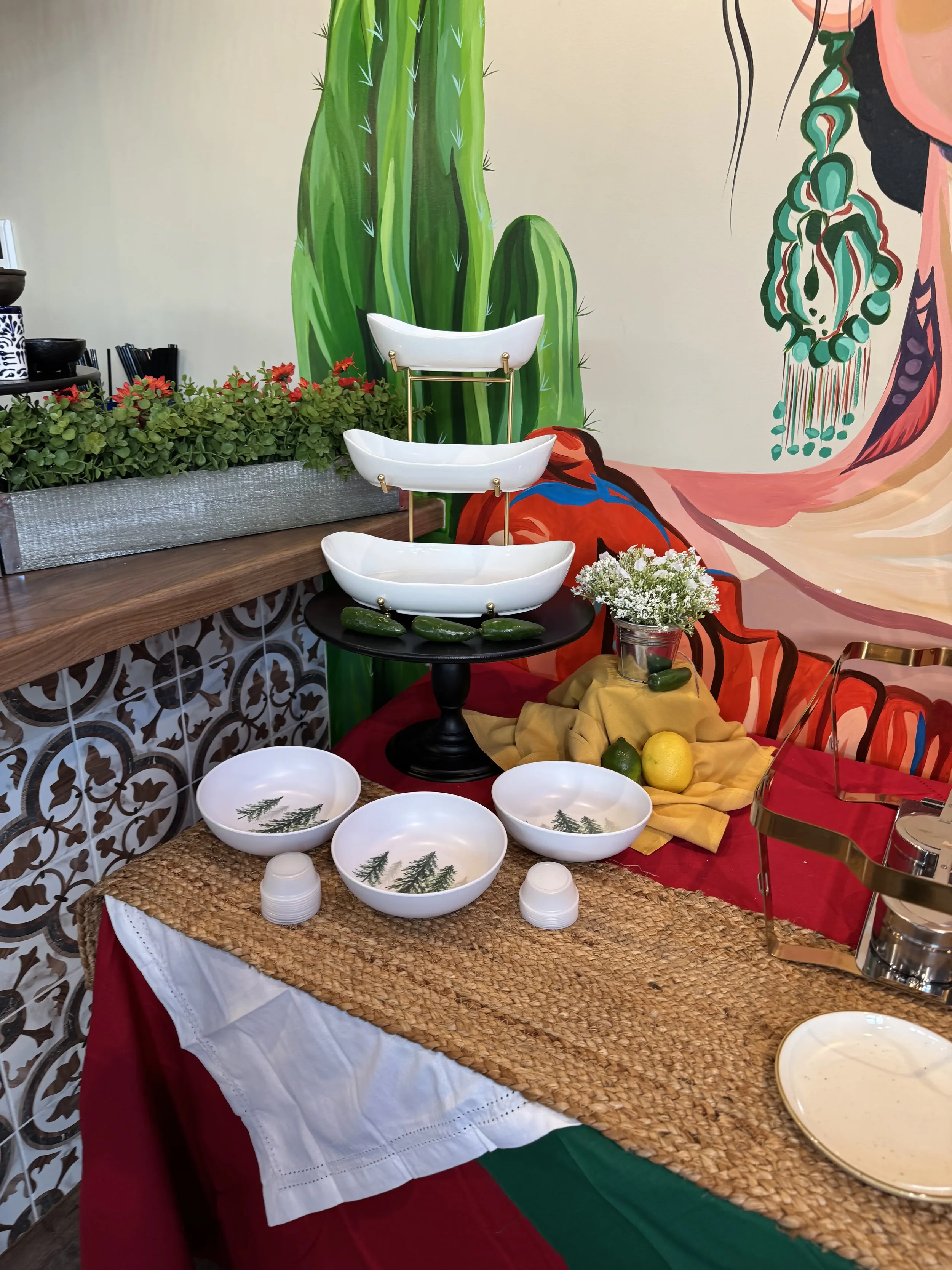Decorative table setting with white bowls featuring green pine tree designs, a black cake stand, a yellow cloth with lemons, and fresh flowers, set against a colorful mural background with cactus and abstract art.