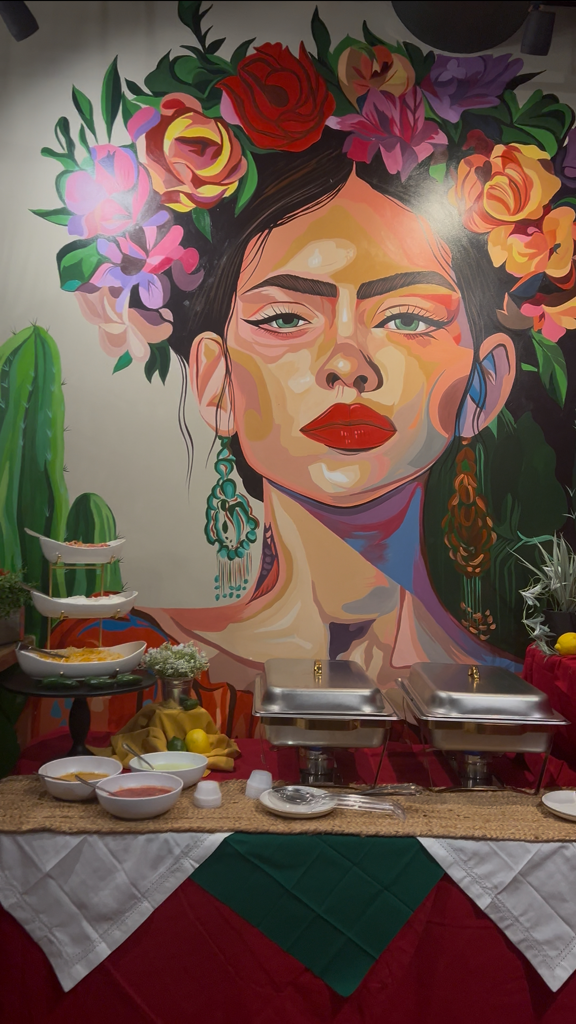 A colorful mural of a woman with floral hair, large earrings, and bold makeup, behind a buffet table with dishes and decorative items, in a vibrant setting.