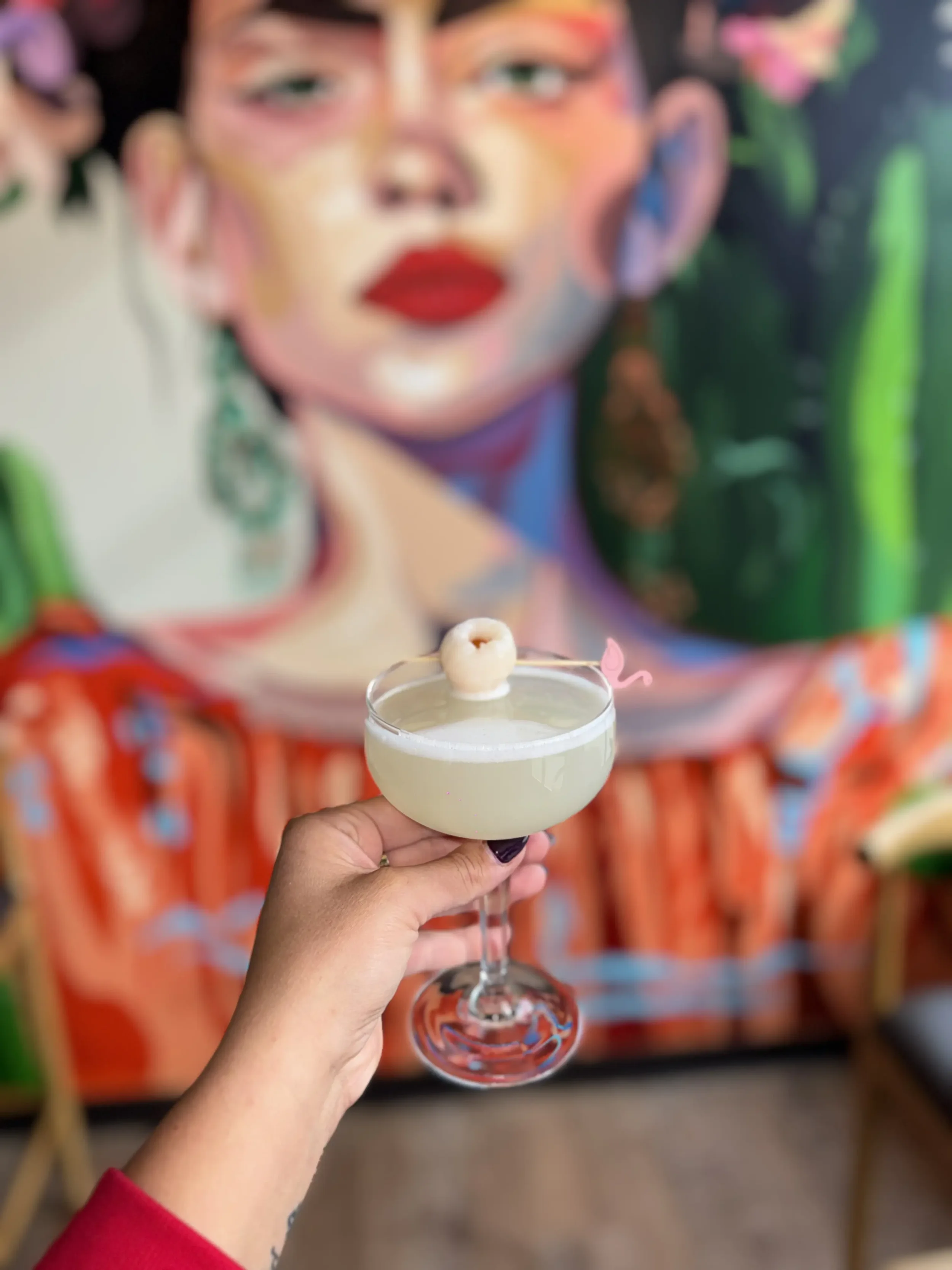 A hand holding a cocktail glass with a light-colored beverage, garnished with a small food item on top, in front of a colorful mural of a woman's face.