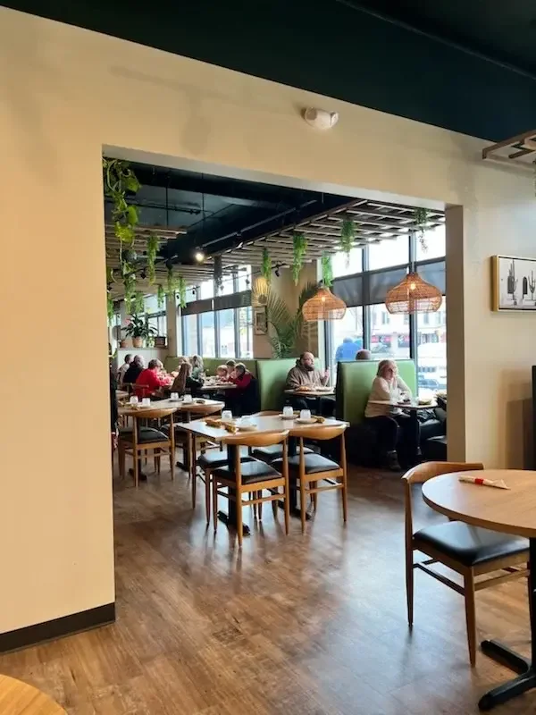 Interior of a modern restaurant with wooden tables, chairs, hanging pendant lights, and large windows letting in natural light; patrons dining and conversing.