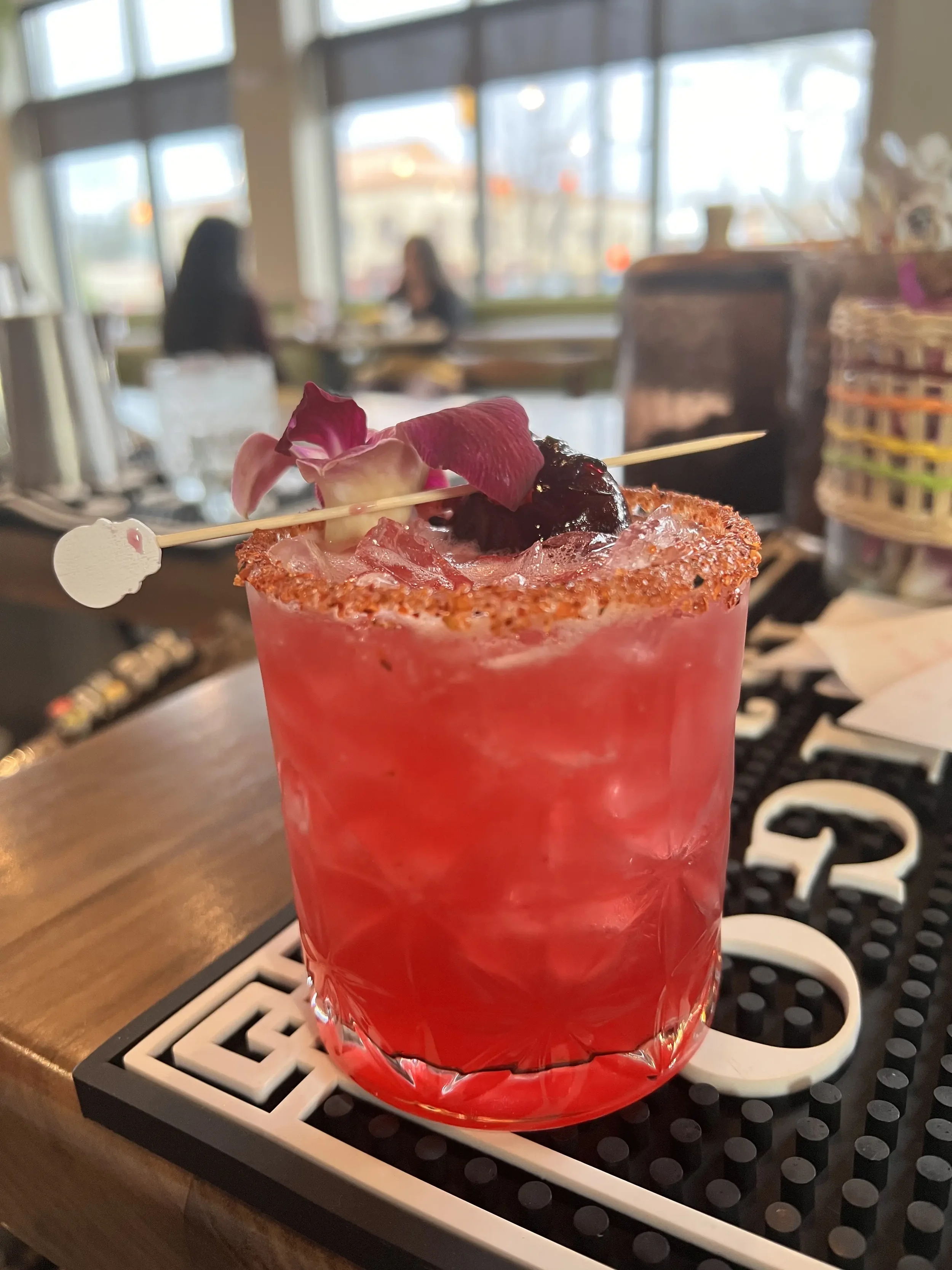 A pink cocktail garnished with flower petals and a cherry, served in a glass with a salted rim, on a bar mat at a restaurant.