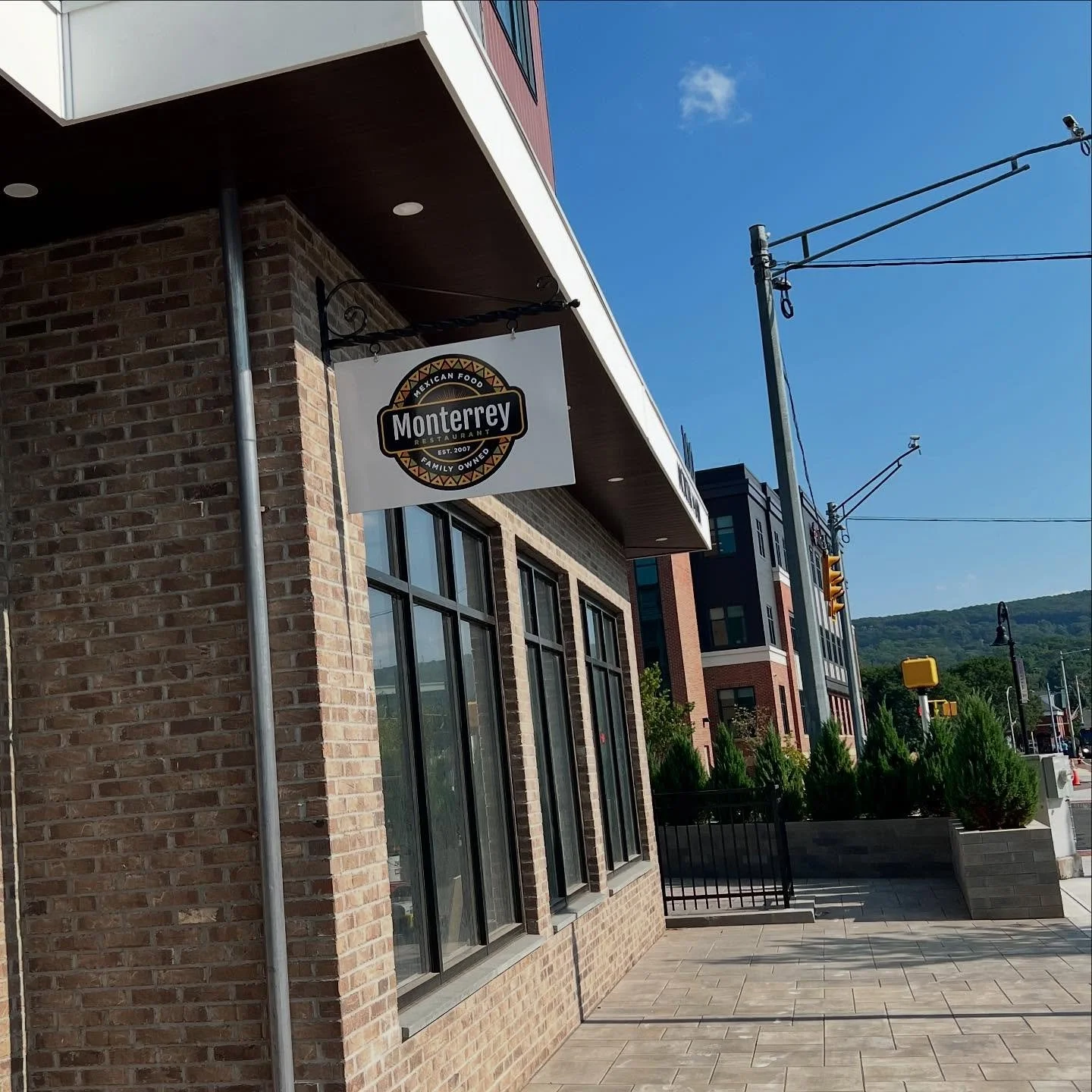 Street view of Monterrey Mexican Food restaurant with a brick exterior, large windows, and a sign hanging above the sidewalk, with modern buildings, power lines, and a mountain in the background.
