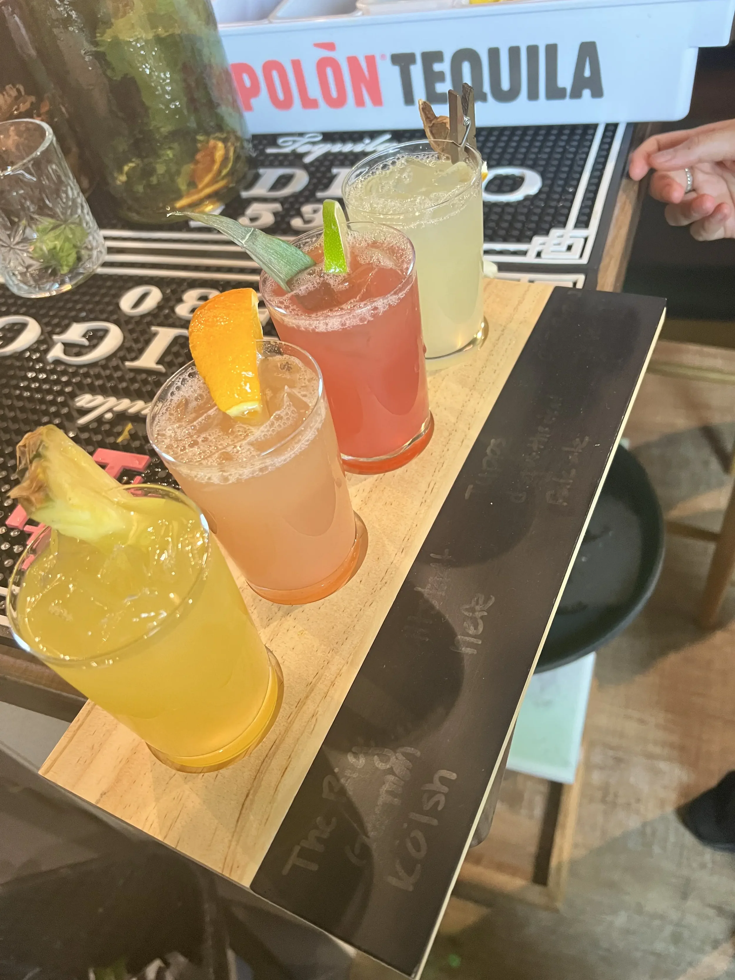 Four colorful cocktails with garnishes on a wooden tray, placed on a table at a bar.