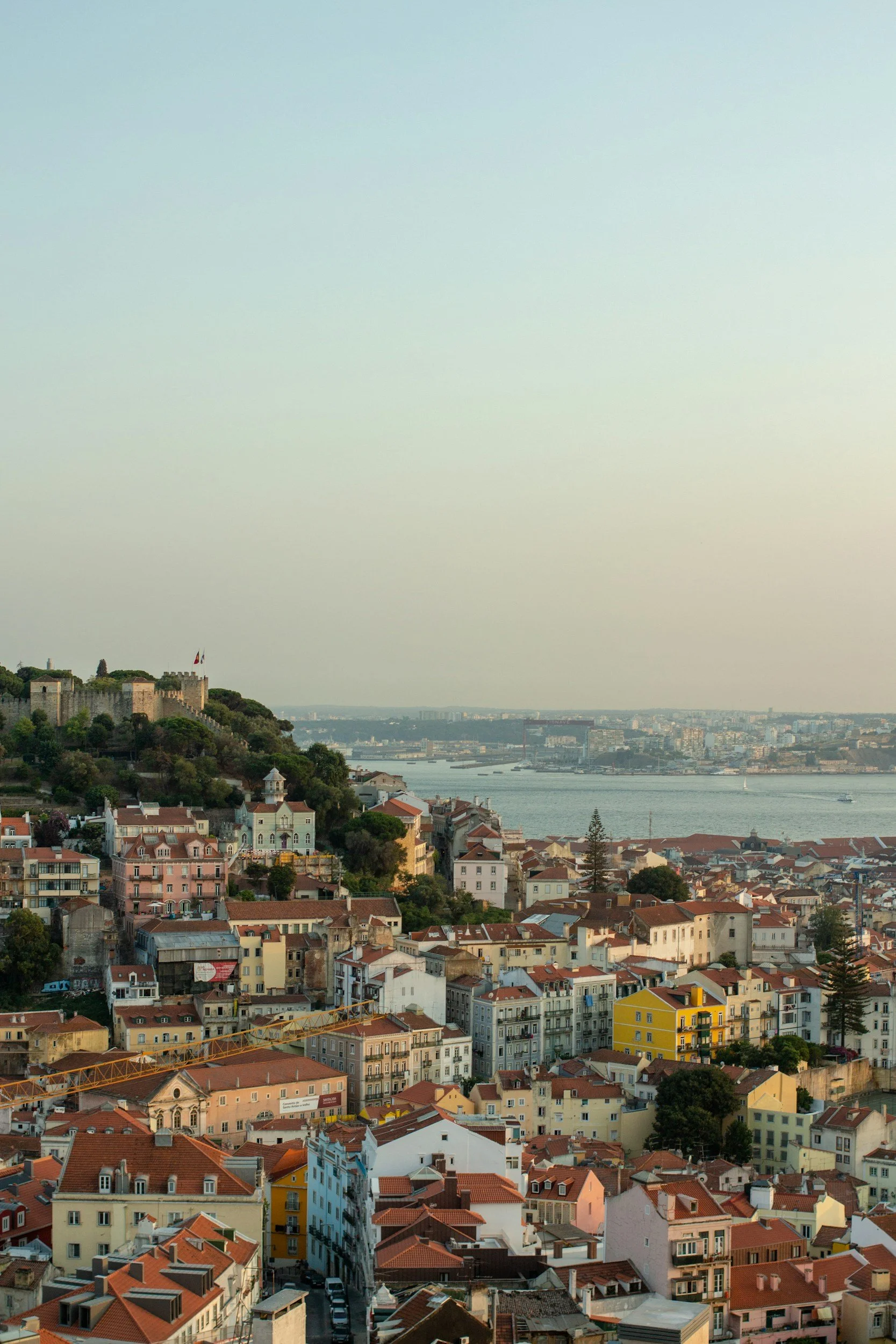 A cityscape showing a hillside with colorful buildings and a fortified structure with flags, overlooking a river with several boats, under a pale sky.