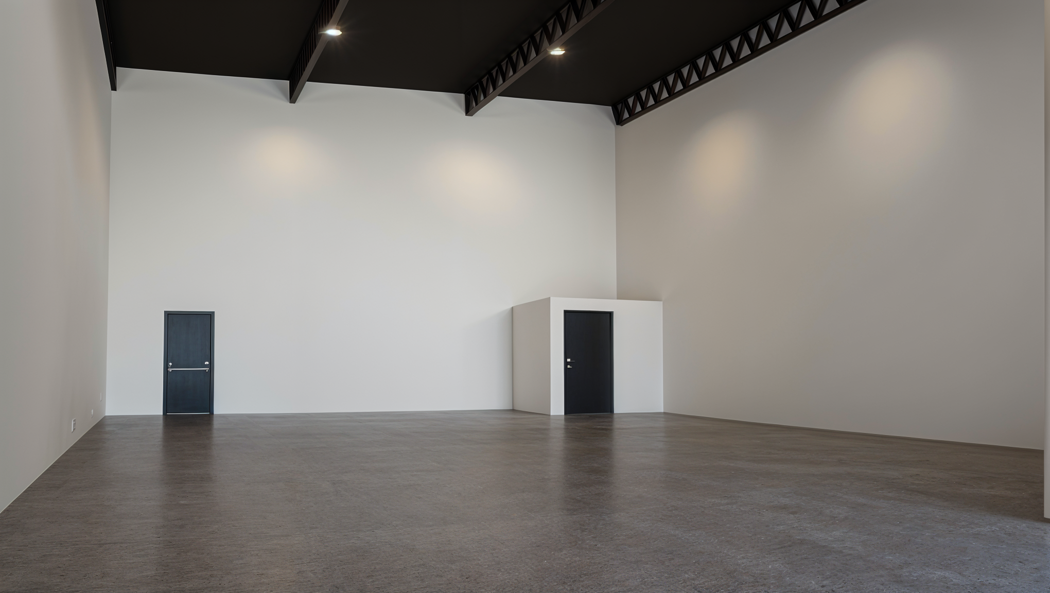Empty gallery room with white and gray walls, wooden floor, black ceiling with ceiling lights, two black doors, one on the left and one in a white enclosure on the right.