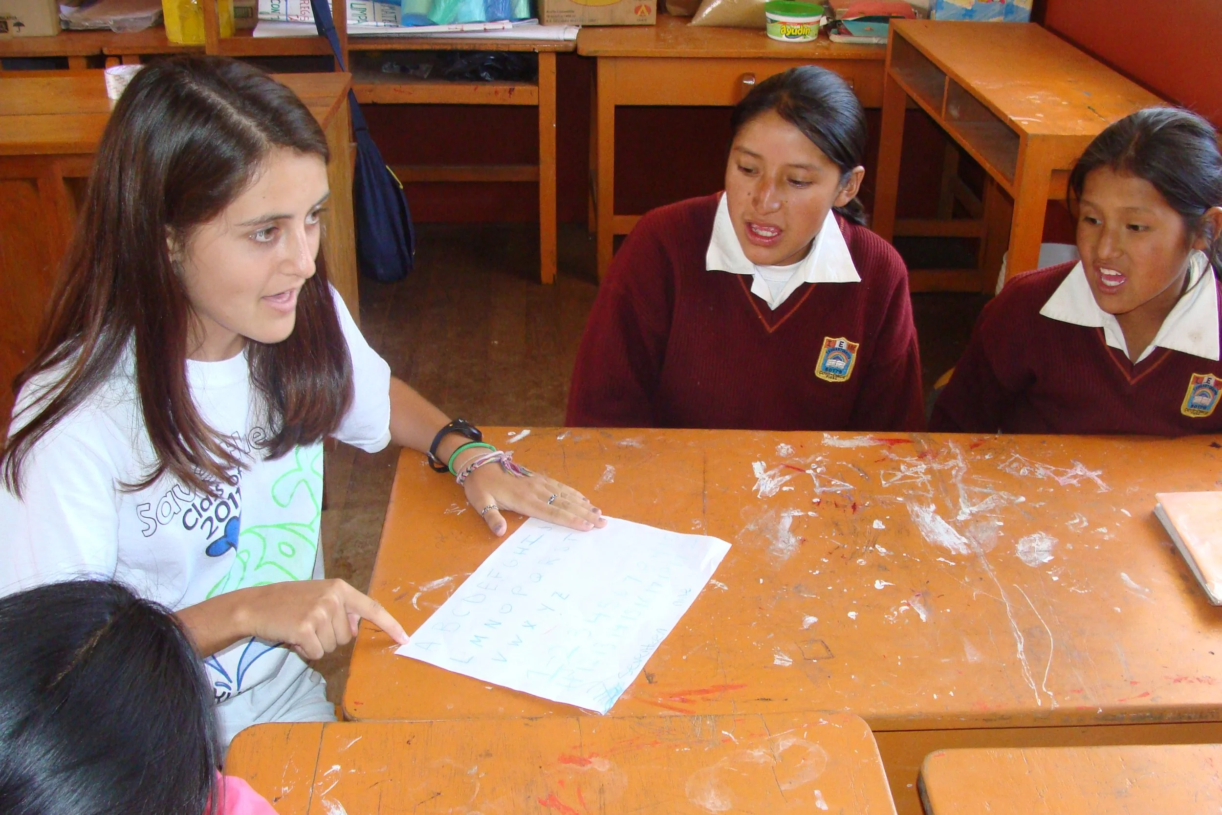 A young woman is sitting at a worn orange classroom desk with three young girls in school uniforms, appearing to explain something written on a piece of paper to them. The classroom has wooden desks and shelves in the background.