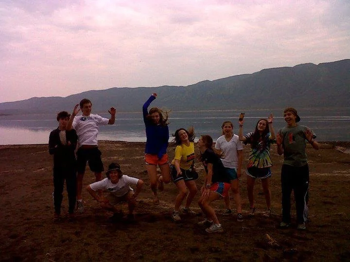 A group of nine young people standing and jumping on a beach near a body of water with mountains in the background, during sunset or dusk.