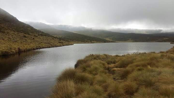 A lake surrounded by grassy hills and mountains, with overcast cloudy sky.
