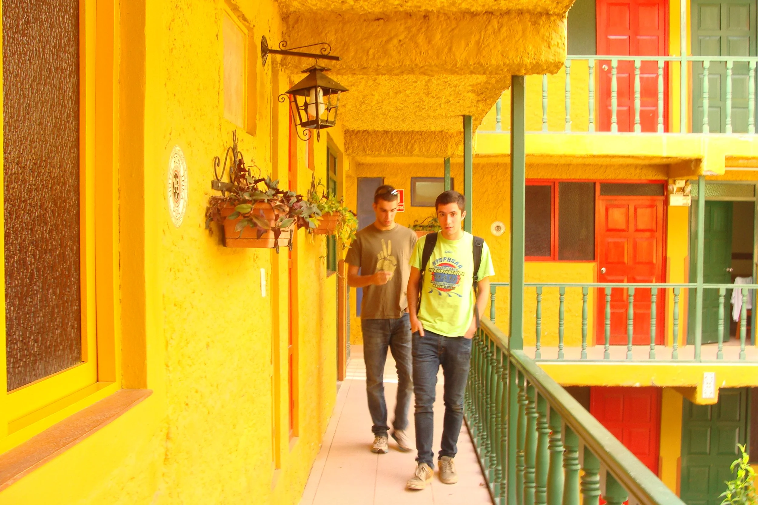 Two young men walking on a narrow outdoor corridor in a colorful yellow building with red and green doors and window shutters.