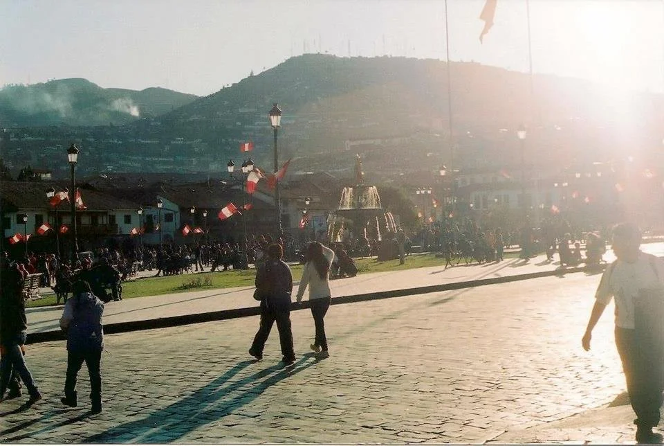 People walking in a town square decorated with multiple French flags, with a fountain in the center, surrounded by buildings and a mountain in the background, during sunset.