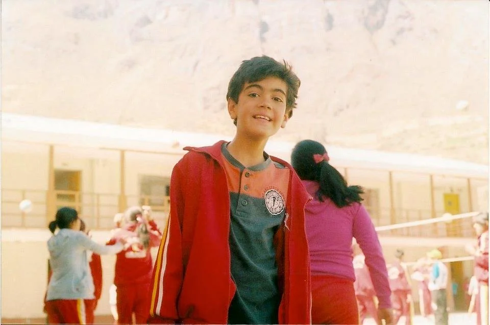 A young boy smiling in front of a crowded school playground with students playing volleyball.