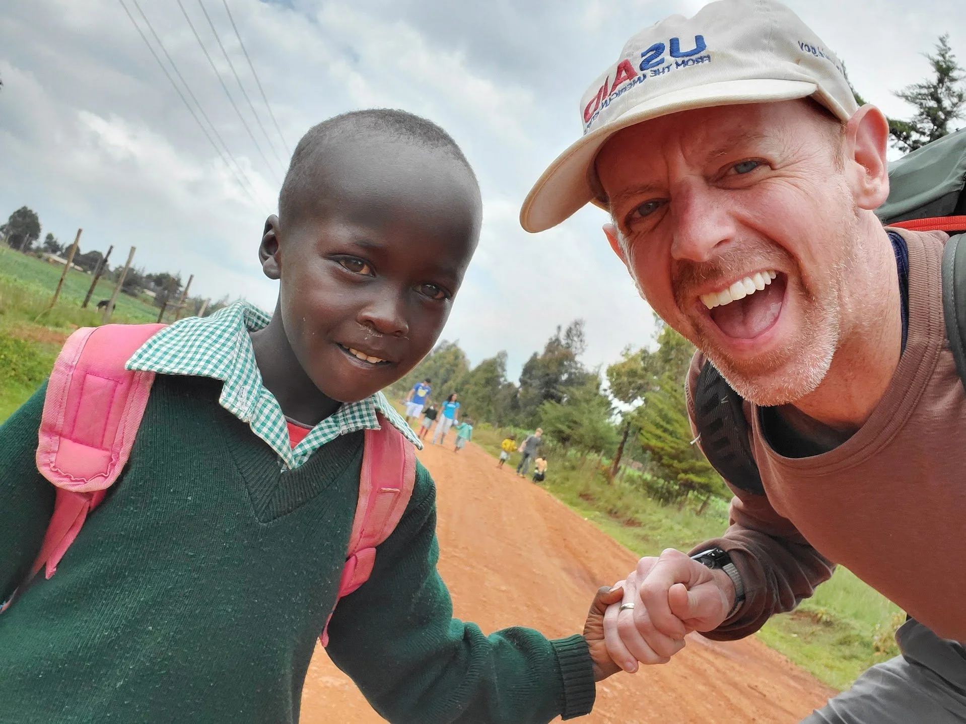 A smiling man in a beige cap shaking hands with a young boy in a green sweater on a dirt road, with a rural landscape in the background.