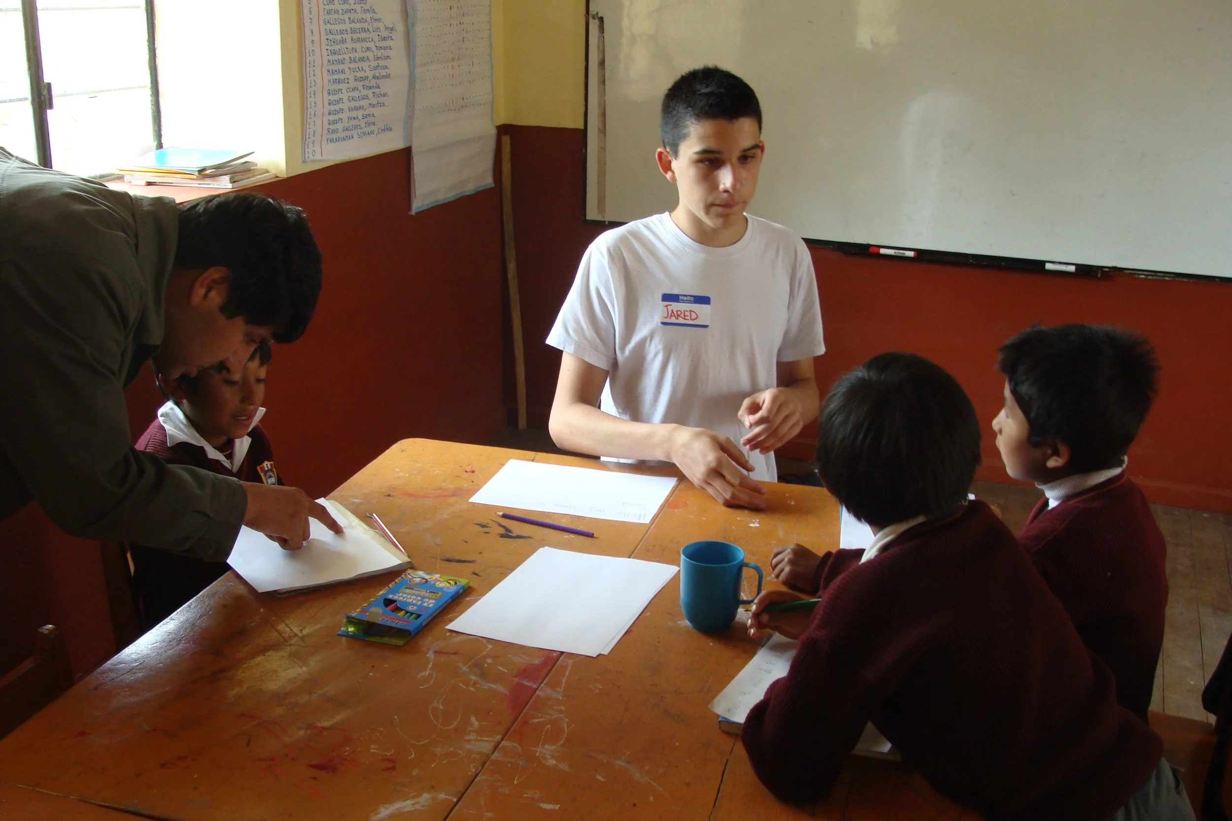 A teacher with a name tag that reads 'Jared' is assisting three young students in a classroom while another adult leans over a student at the table. The students are focused on their work, and there are papers, a blue mug, and colorful markers on the