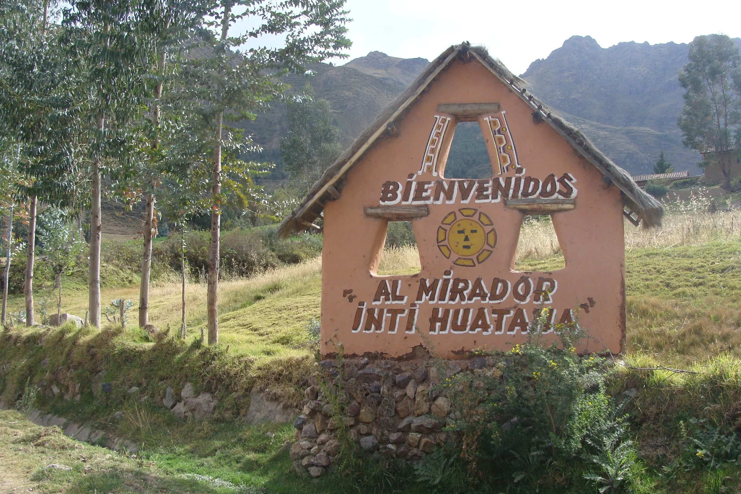 Welcome sign in a rural area with mountains in the background, featuring a thatched roof and painted text in Spanish.