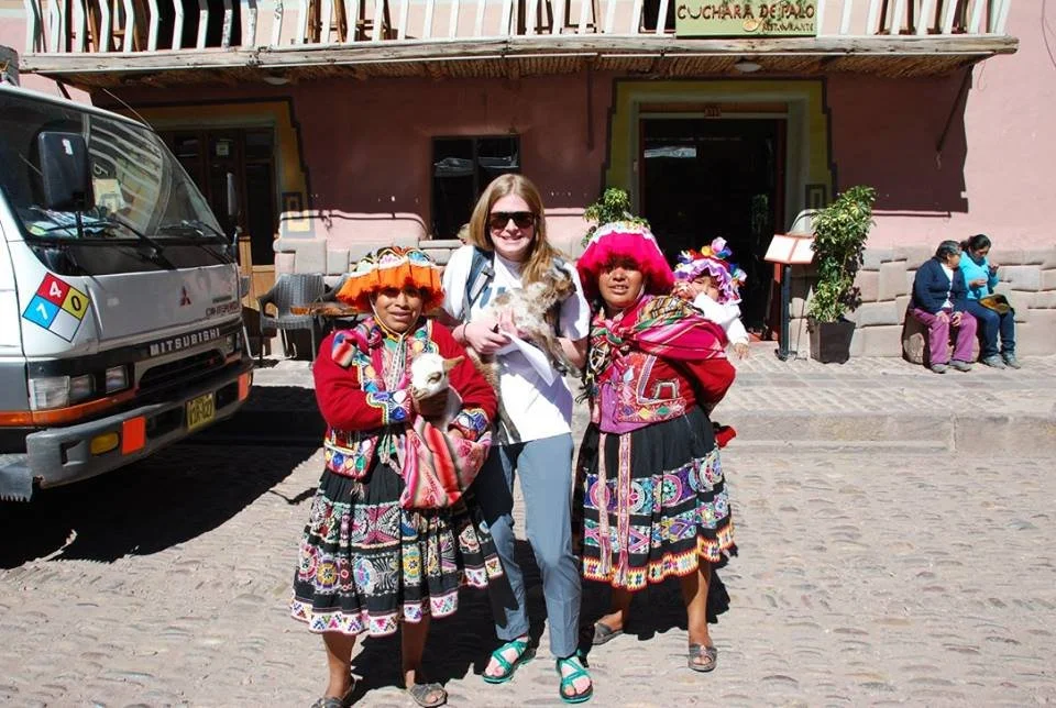 A woman standing between two indigenous women in traditional colorful clothing on a cobblestone street in front of a pink building. The woman wears sunglasses, a white shirt, and jeans. Two people sit on a bench in the background.