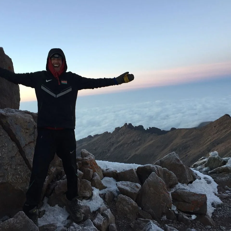 A person dressed in winter outdoor gear standing on rocky terrain at high elevation above clouds at sunset or sunrise, with arms outstretched and smiling.