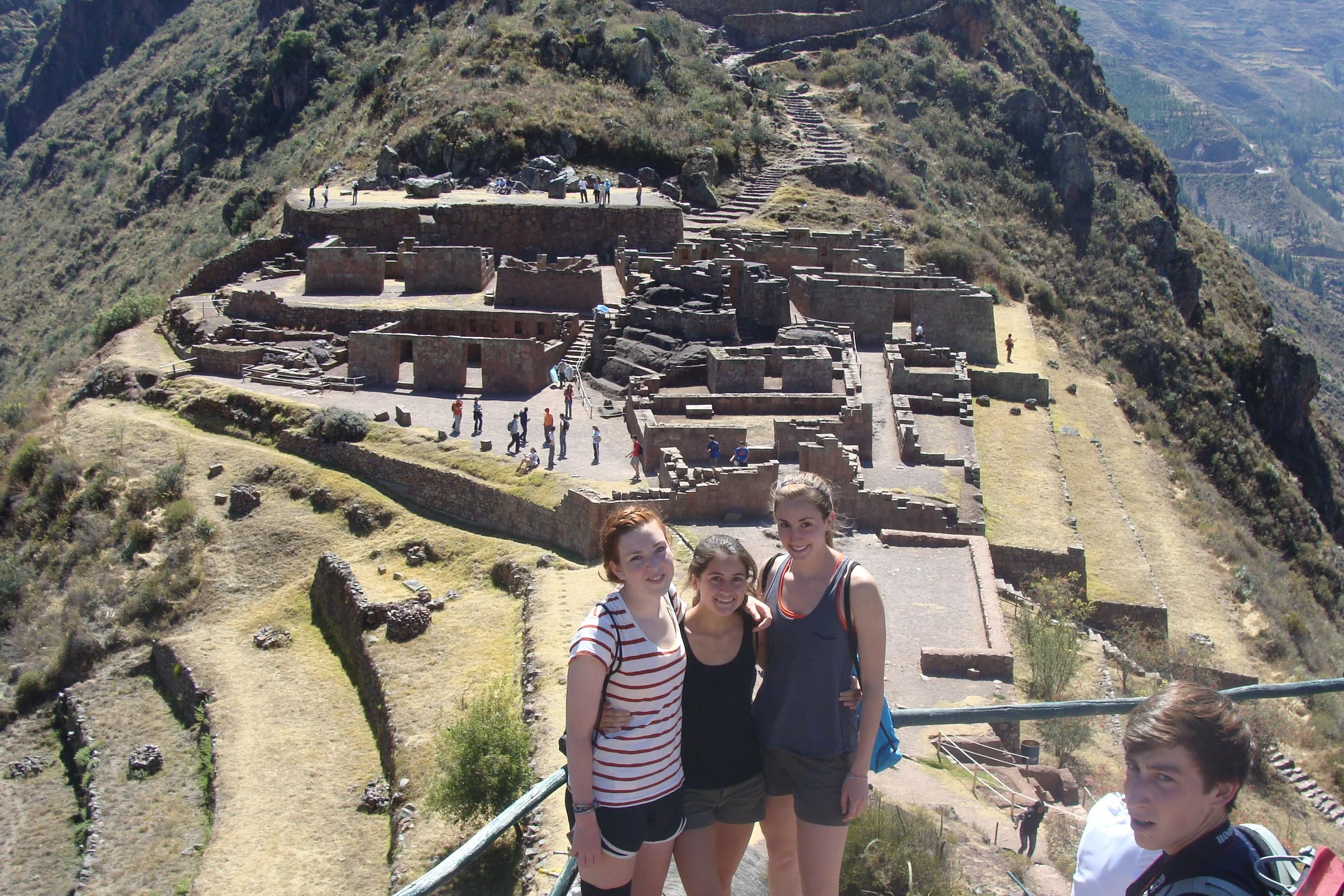 Three young women and a young man standing together outdoors near ancient Inca ruins on a hilltop with the Machu Picchu archaeological site visible in the background.