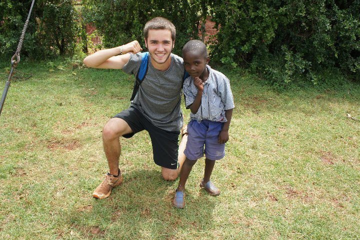 A young man kneeling on one knee next to a young boy in a park, both smiling and posing for the camera.